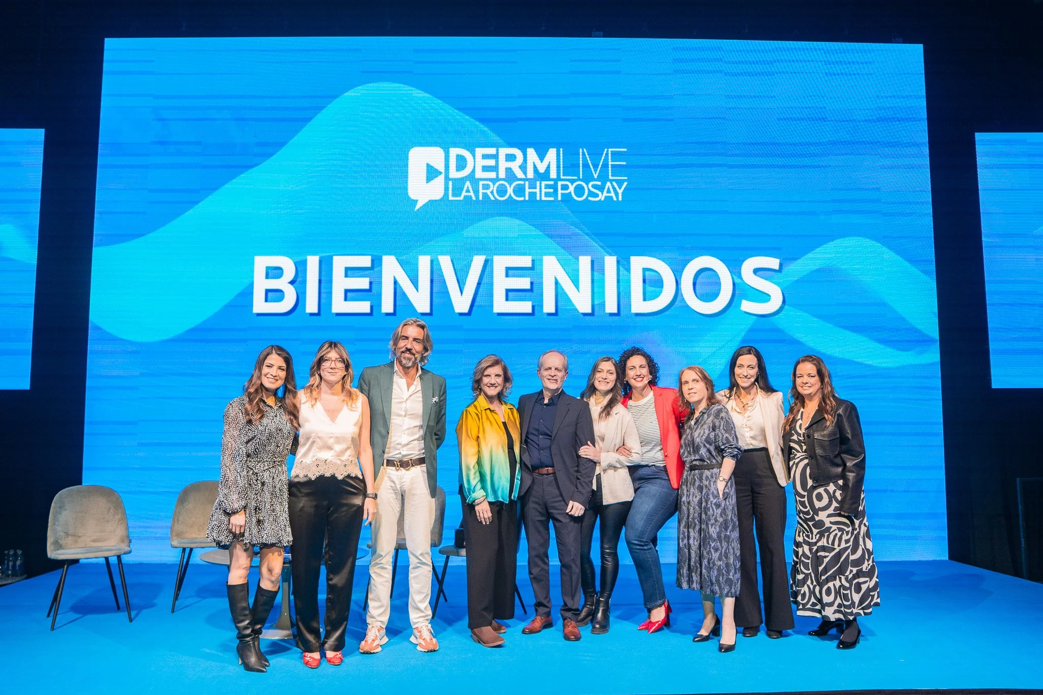 Grupo de personas posando en un escenario con una pantalla grande de fondo que dice 'BIENVENIDOS', en un evento de Derm Live La Roche Posay.