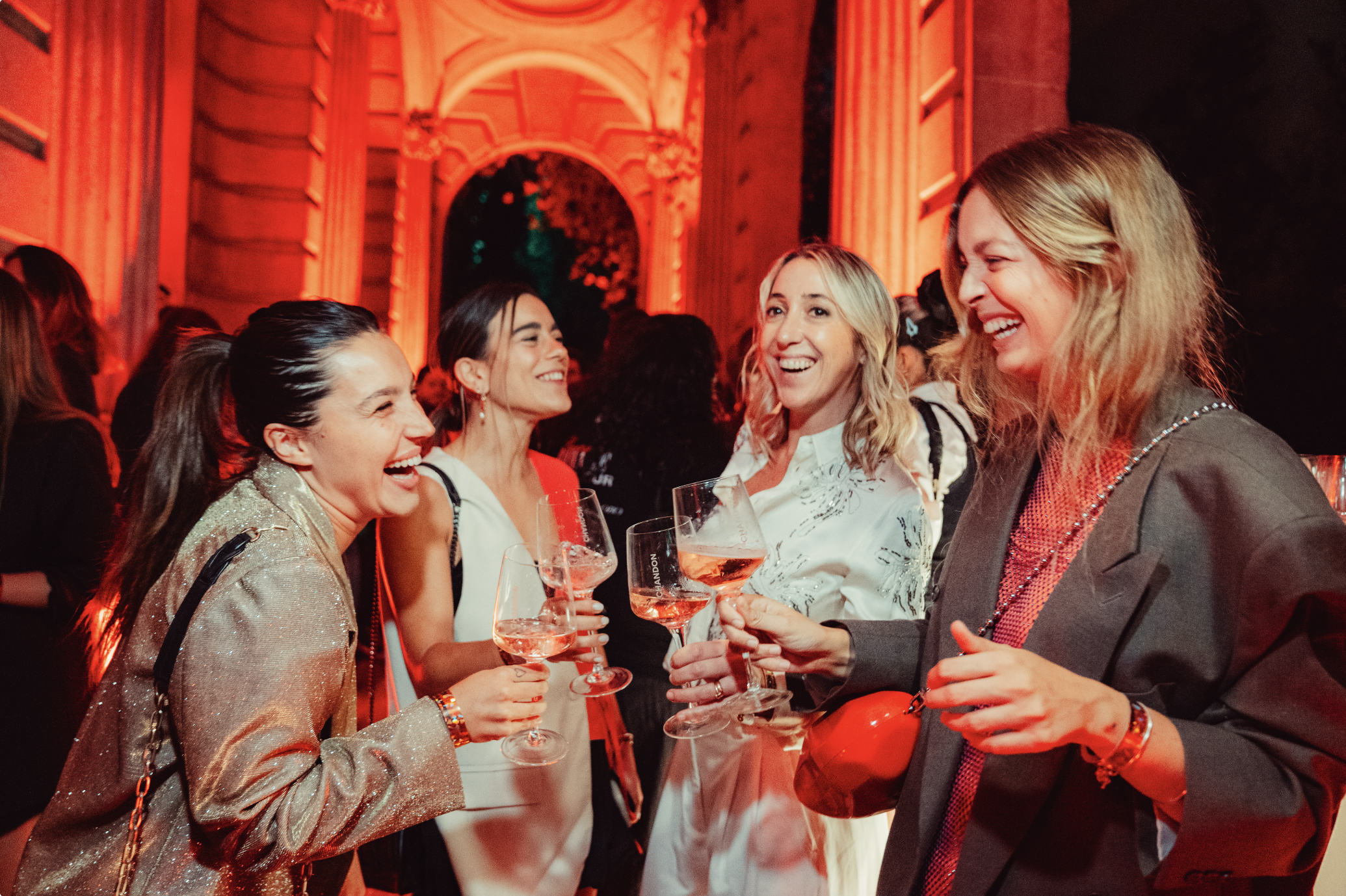 Grupo de mujeres sonriendo y brindando con copas de vino en una fiesta en un recinto con iluminación cálida y decoraciones elegantes.