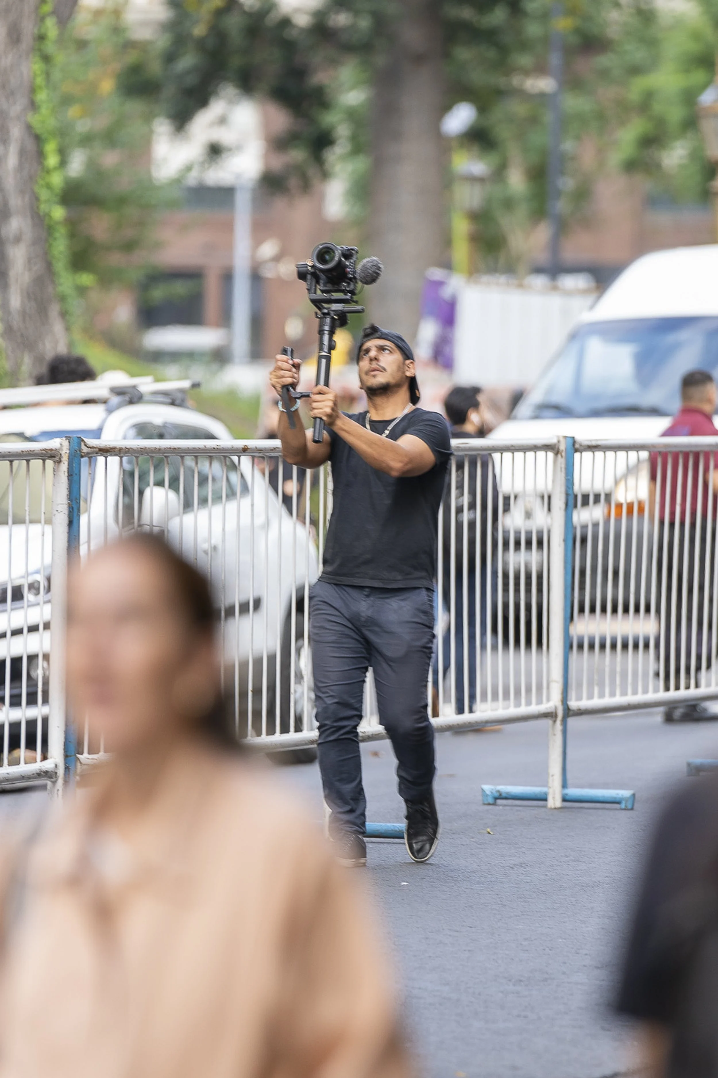Un hombre con gorra ajusta una cámara de video en medio de una calle, con una cerca metálica frente a él y varias personas en el fondo, en un entorno urbano con árboles y vehículos.