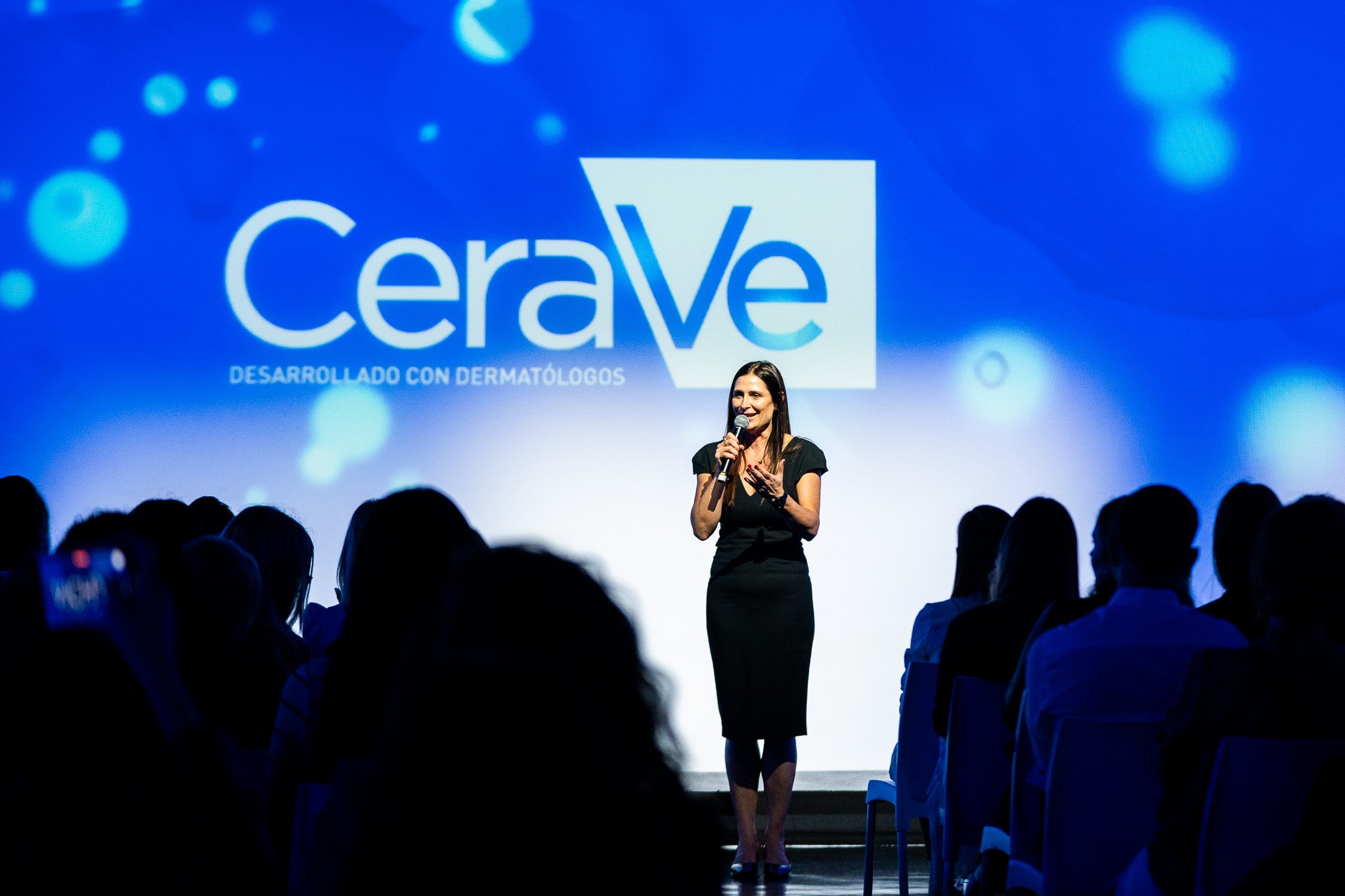 Una mujer habla frente a una audiencia en un escenario con un proyector que dice 'Cerave desarrollada con dermatólogos'.