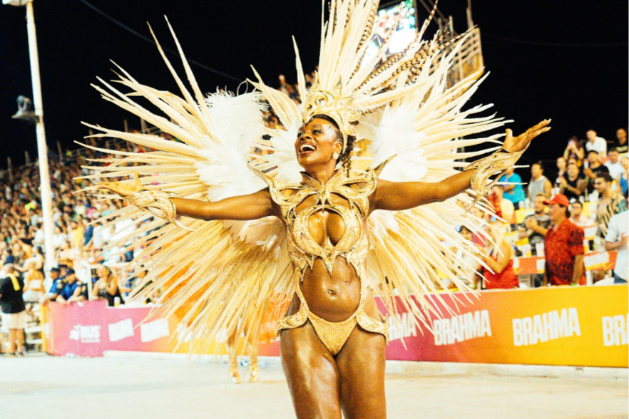 Mujer en disfraz colorido y elaborado, con plumas grandes, en un carnaval, rodeada de espectadores.