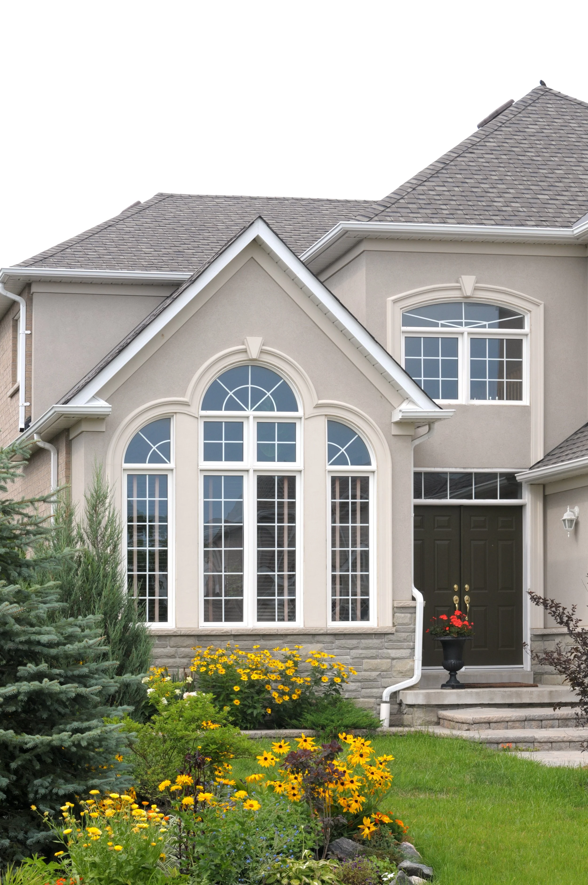 Front of a house with large arched windows, surrounded by a garden with yellow flowers, shrubs, and a small pine tree.