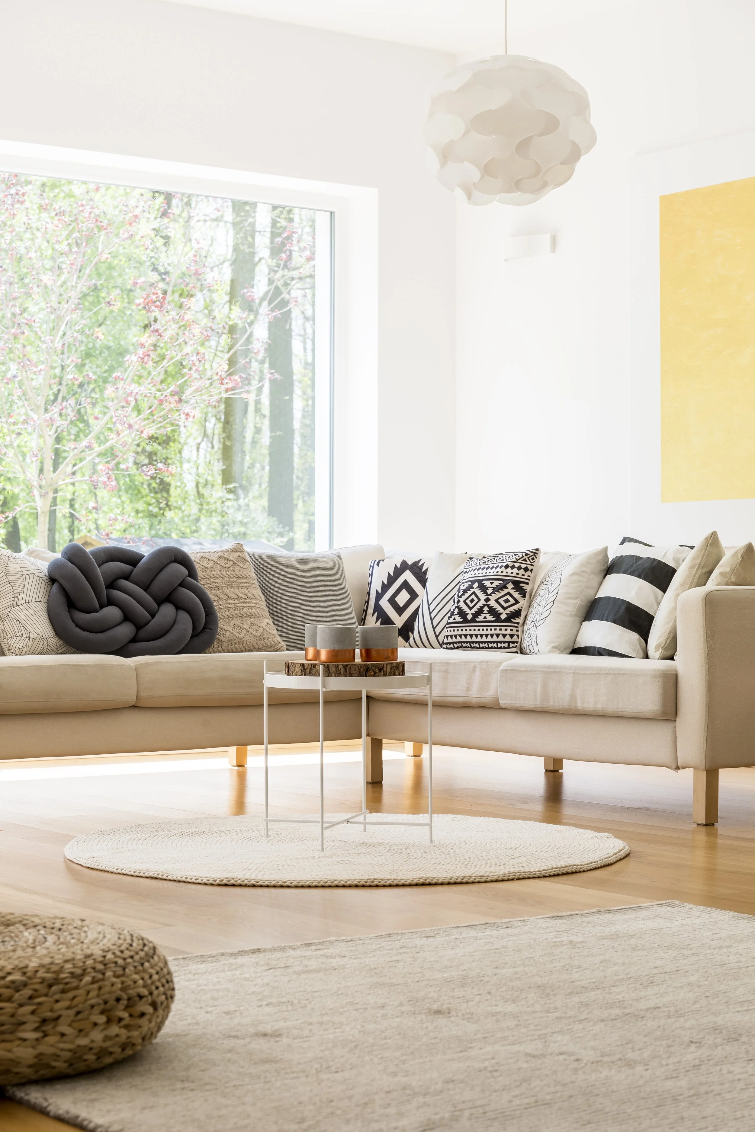 Bright living room with beige sectional sofa, black and white patterned pillows, a small round white side table, and a woven pouf on a cream rug. Large window shows trees outside, and a white modern pendant light hangs from the ceiling.
