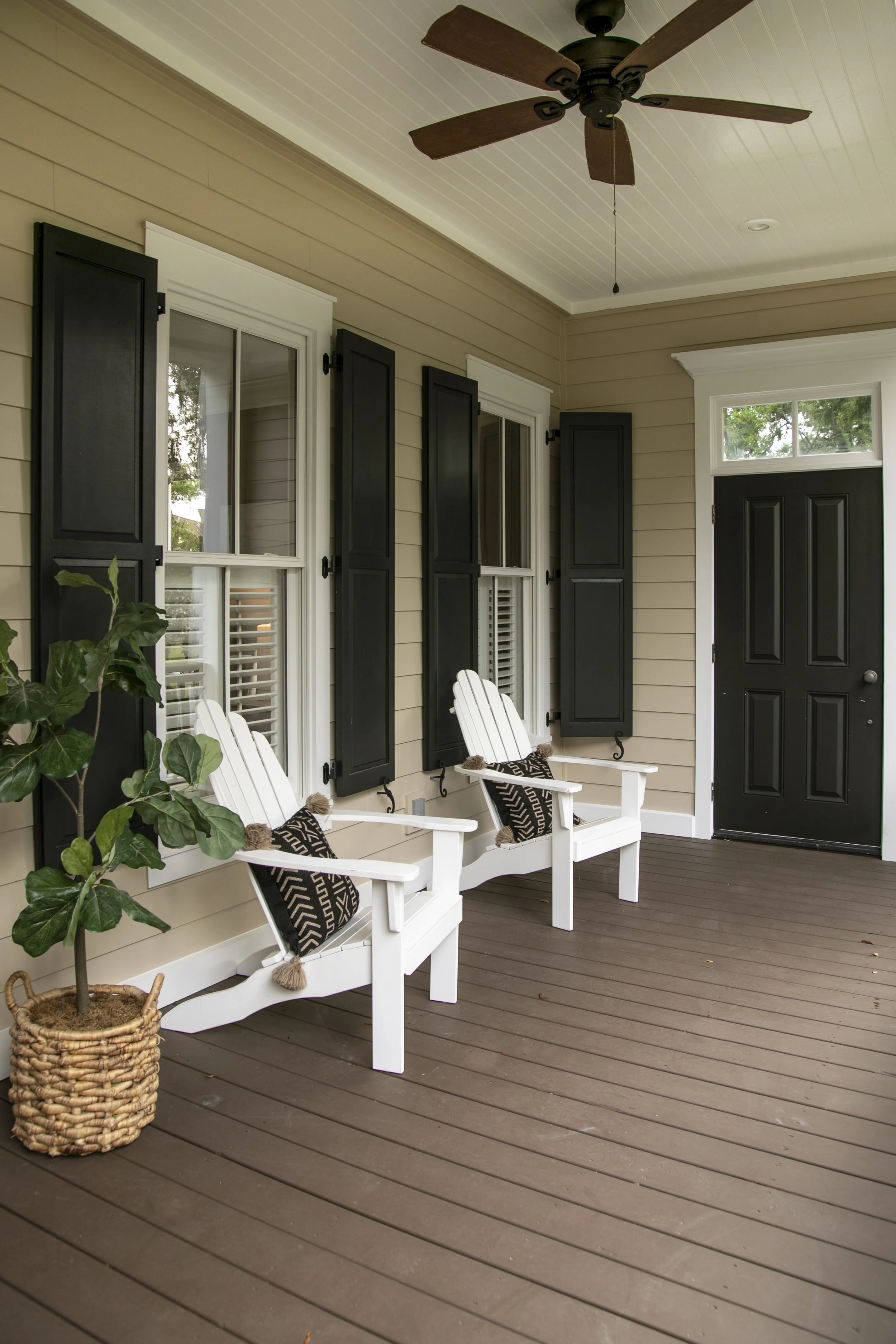 Front porch with beige siding, white trim, black window shutters, black front door with sidelights, two white Adirondack chairs with patterned pillows, potted plant, and ceiling fan, with a wooden deck floor.