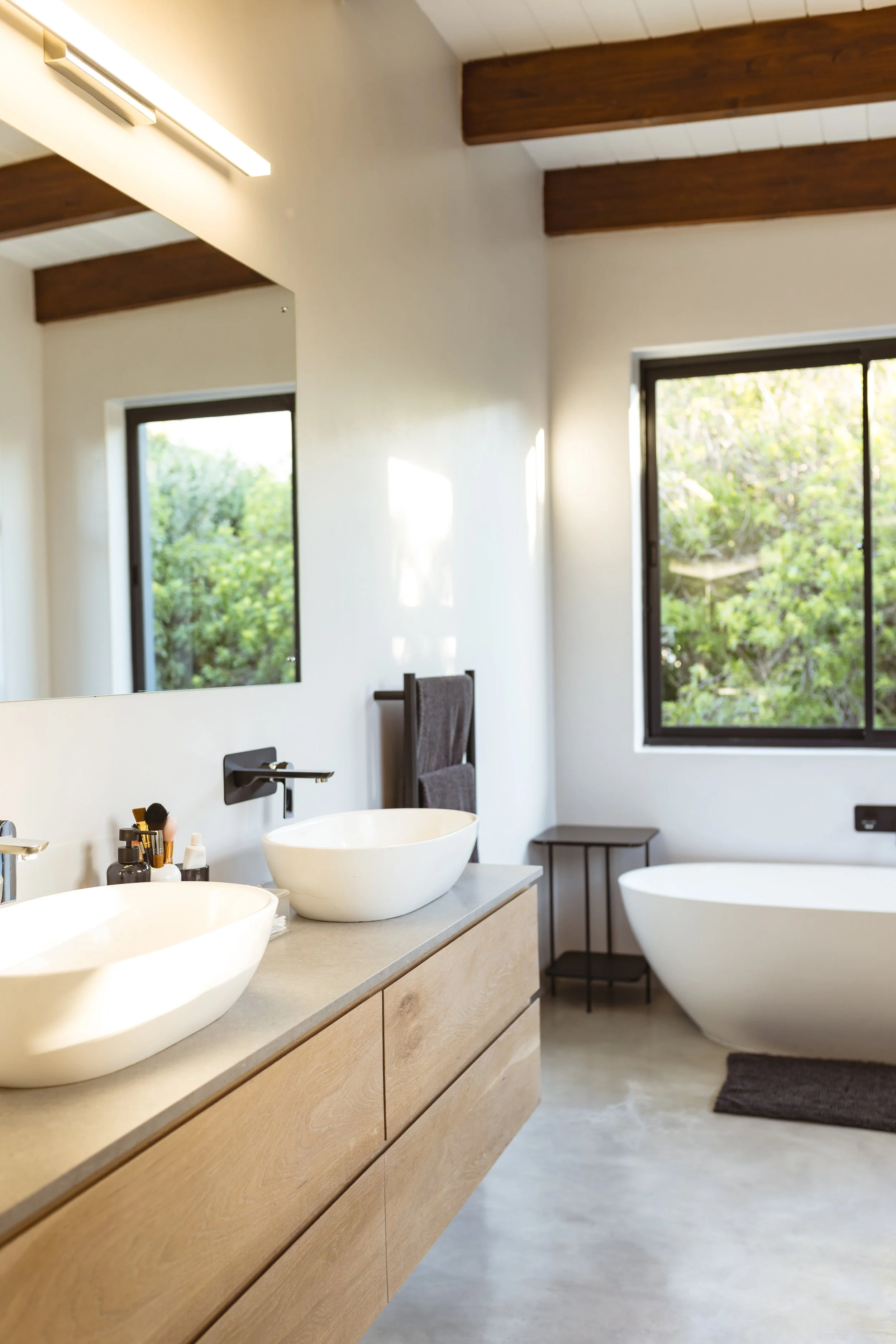 Modern bathroom with a double vanity with round vessel sinks, a large mirror, black wall-mounted faucets, and a bathtub with wooden beams on the ceiling and large windows with greenery outside.