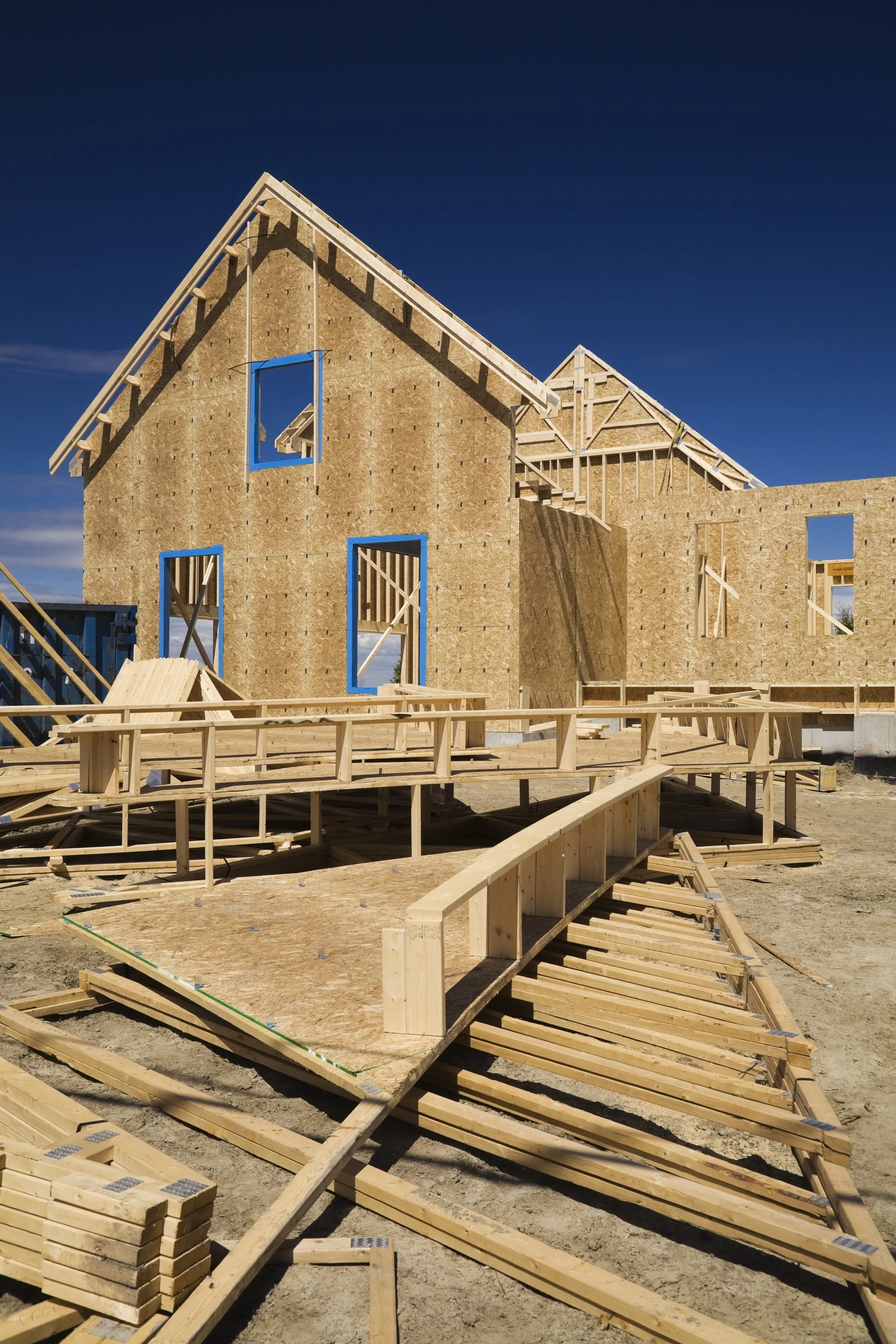 A house under construction with wooden framing and plywood sheathing on the walls, showing open window and door spaces, and assembled roof trusses, against a clear blue sky.