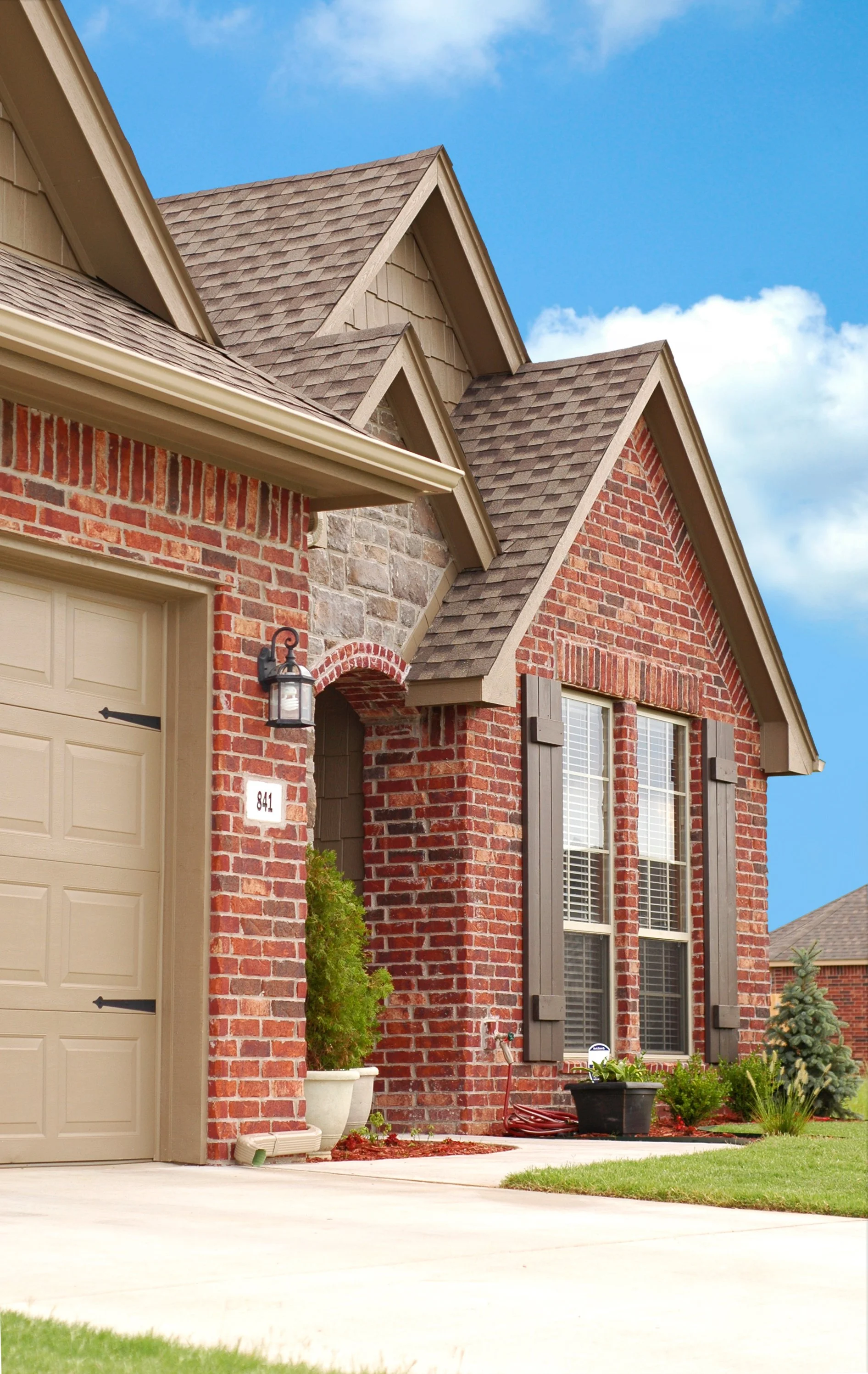 Front view of a brick house with a beige garage door, black shutters, and a gable roof with brown shingles under a partly cloudy blue sky.