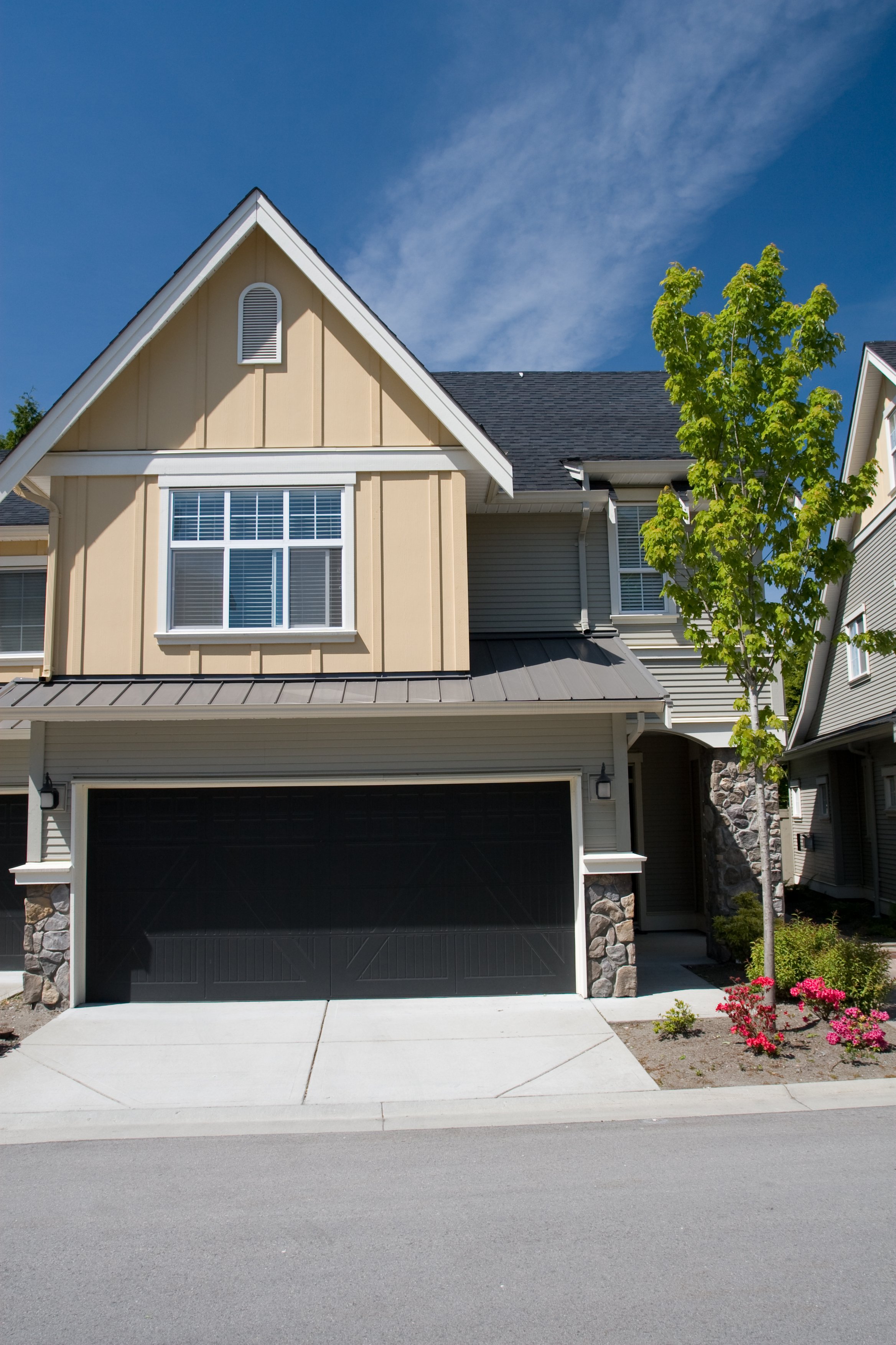 Front view of a modern house with beige and gray siding, a dark garage door, a small tree, and pink flowers in the landscaped yard under a blue sky.