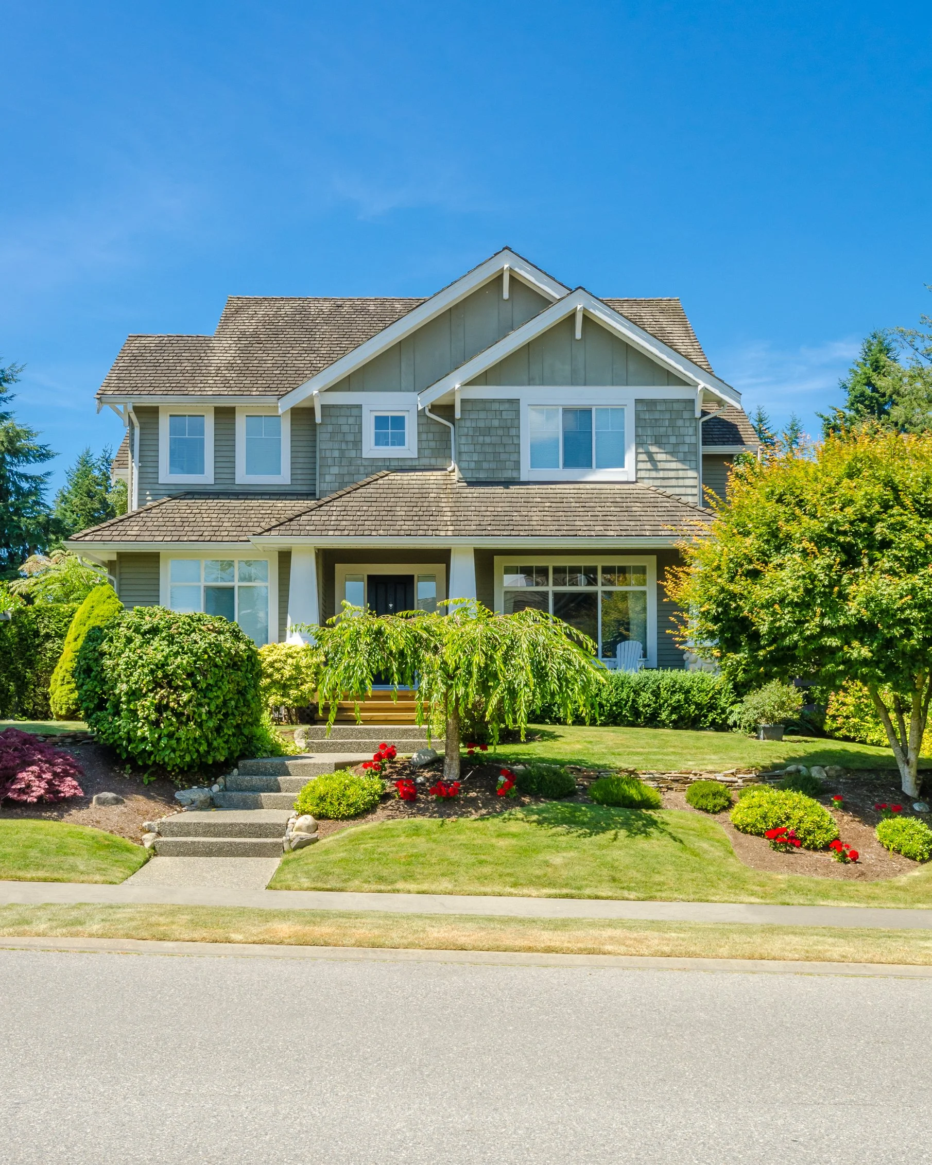 Front view of a two-story house with a brown shingle roof, gray siding, and multiple windows. There is a front yard with a painted concrete pathway leading to the front door, surrounded by green plants, bushes, and some red flowers. The sky is clear and blue.