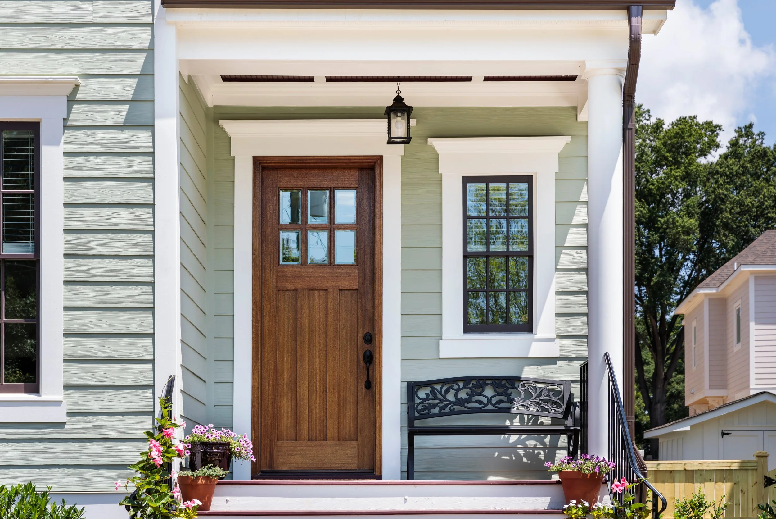 Front porch of a house with light green siding, a wooden front door with window panes, black lantern-style light fixture, and black metal bench. Potted flowers decorate the steps, and the house has white trim and columns. Trees are visible in the background under a partly cloudy sky.