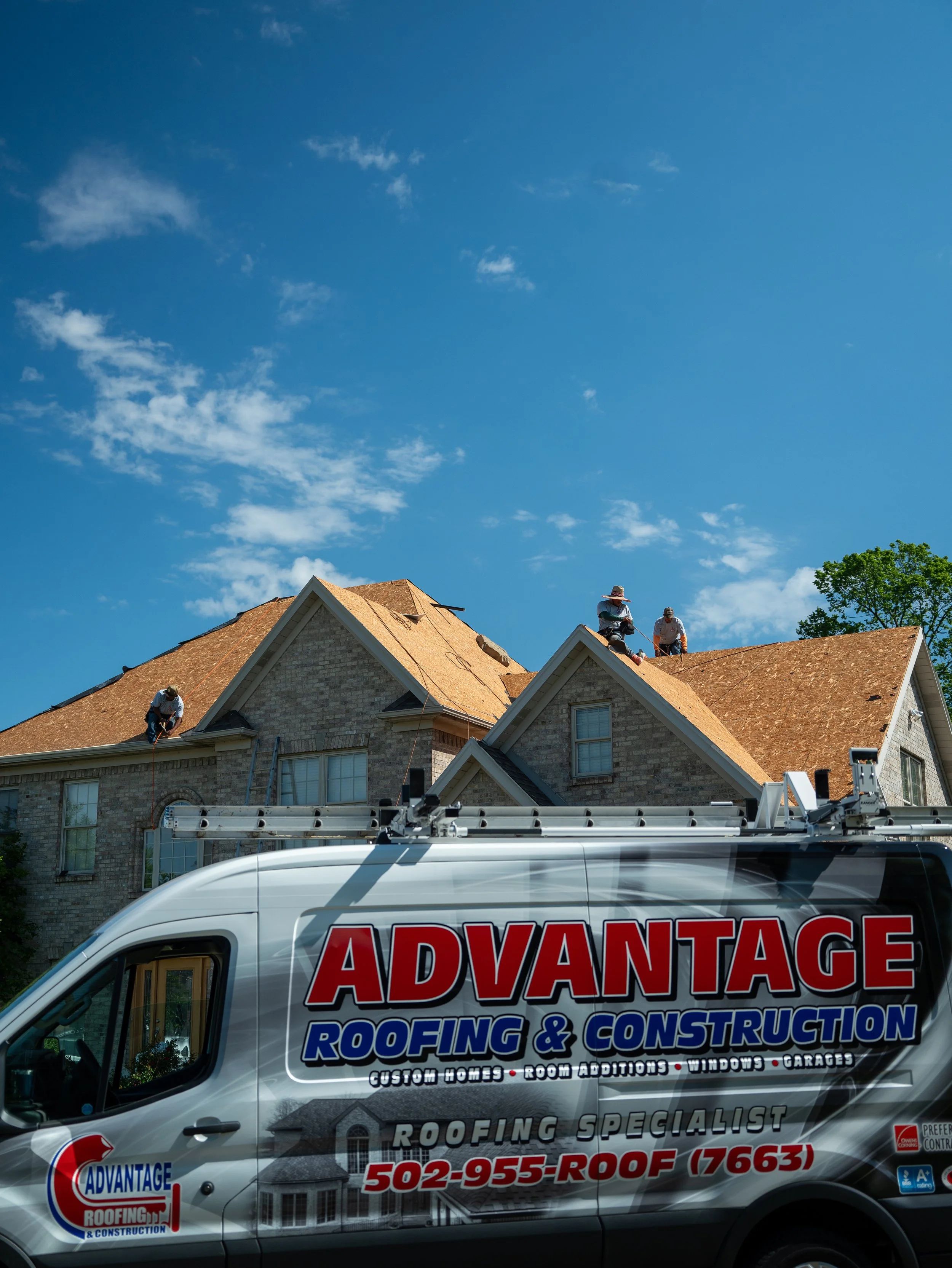 Construction workers inspecting a roof on a residential house under a blue sky with a few clouds. A company van with roofing and construction services is parked in front.