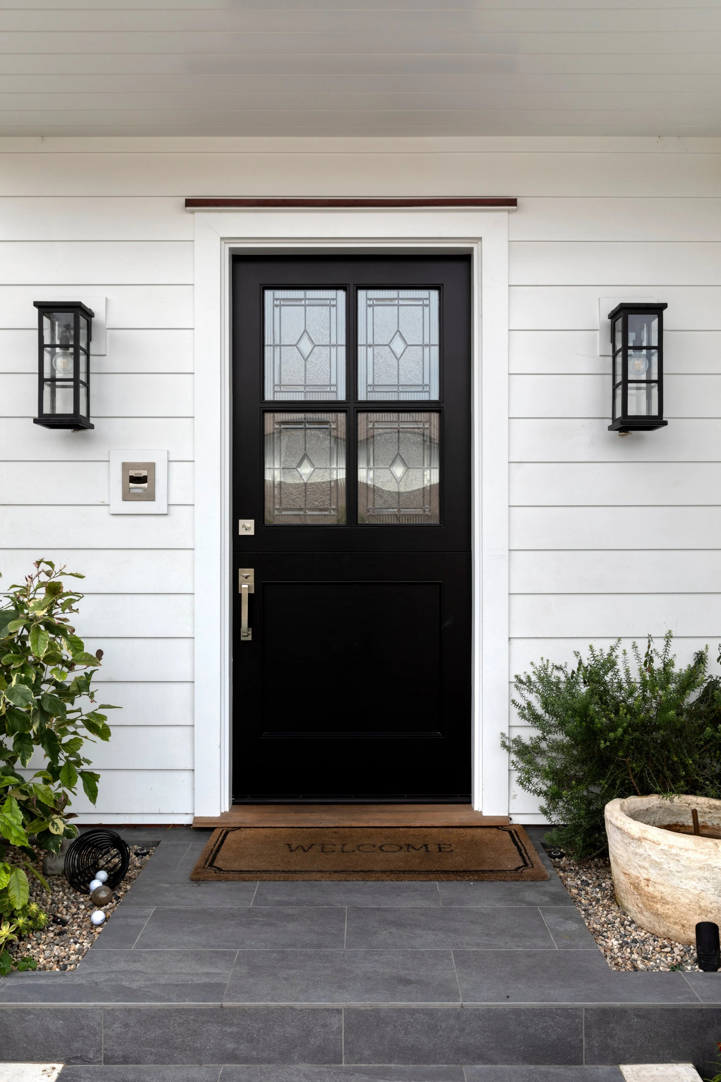Front porch with black front door featuring glass panels, white siding, wall-mounted black lantern lights, welcome mat, potted plants, and decorative gravel.