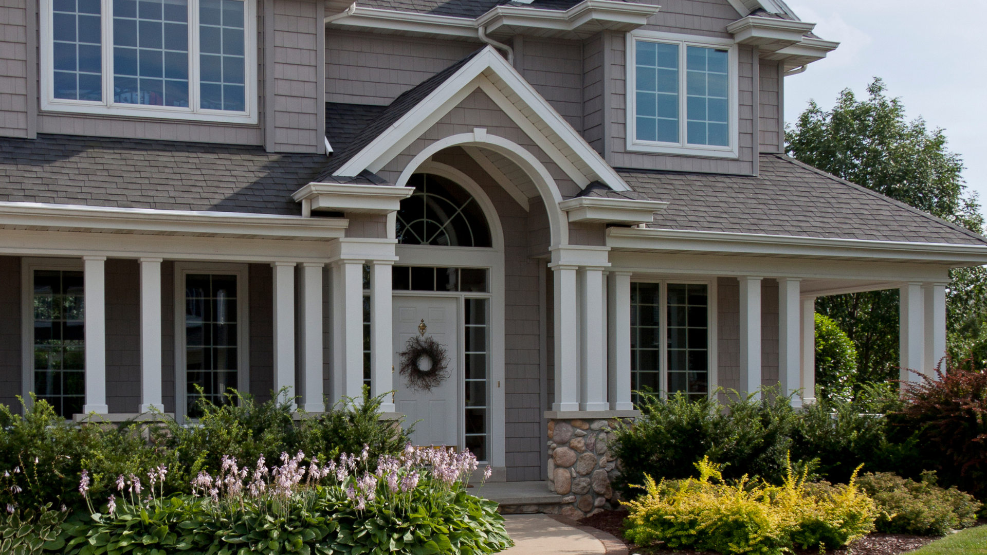 Front view of a two-story house with beige siding, white trim, and a gabled roof, surrounded by green shrubs and flowering plants.