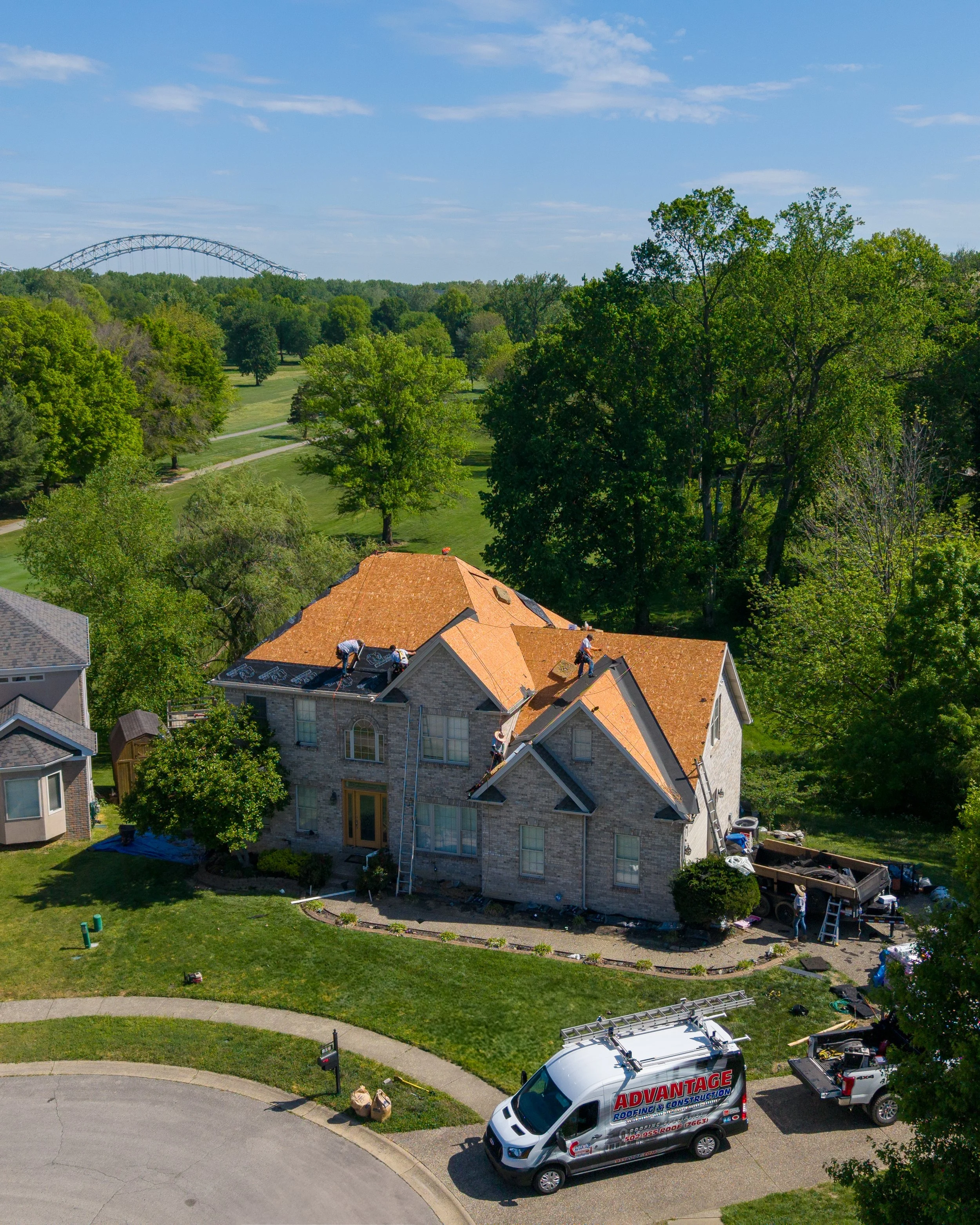 A house with a roof under construction, workers on the roof, a ladder, and construction vehicles nearby, including a truck labeled 'Advantage Roofing & Construction'. Trees and a clear blue sky are in the background.