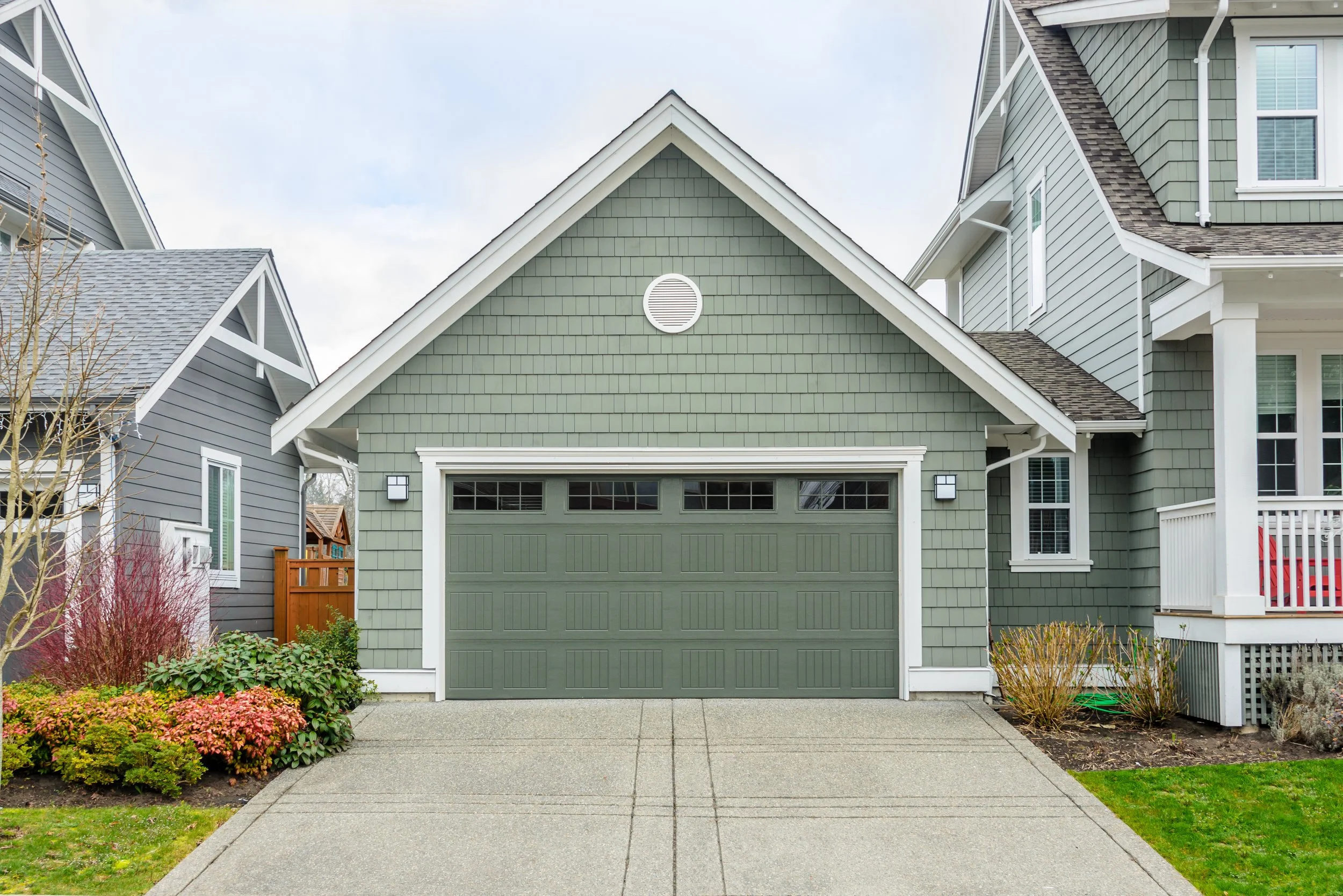 Front view of a green house with a garage door, surrounded by landscaping, and neighboring houses on each side.