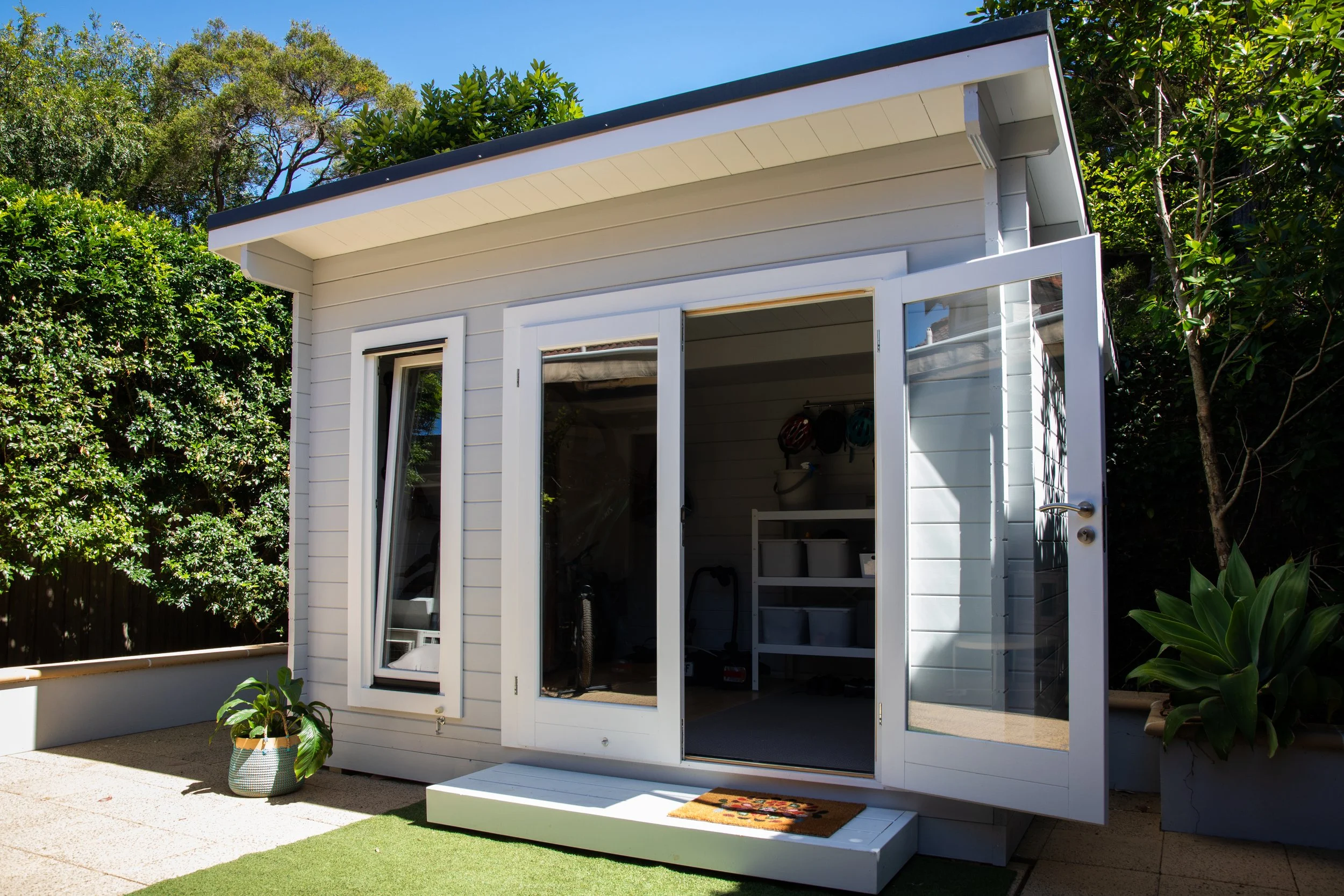 A small white shed with sliding glass doors is set in a backyard with green trees and bushes. The shed has a dark trim and a sloped roof. A potted plant sits outside on a concrete surface next to a small shelf inside.