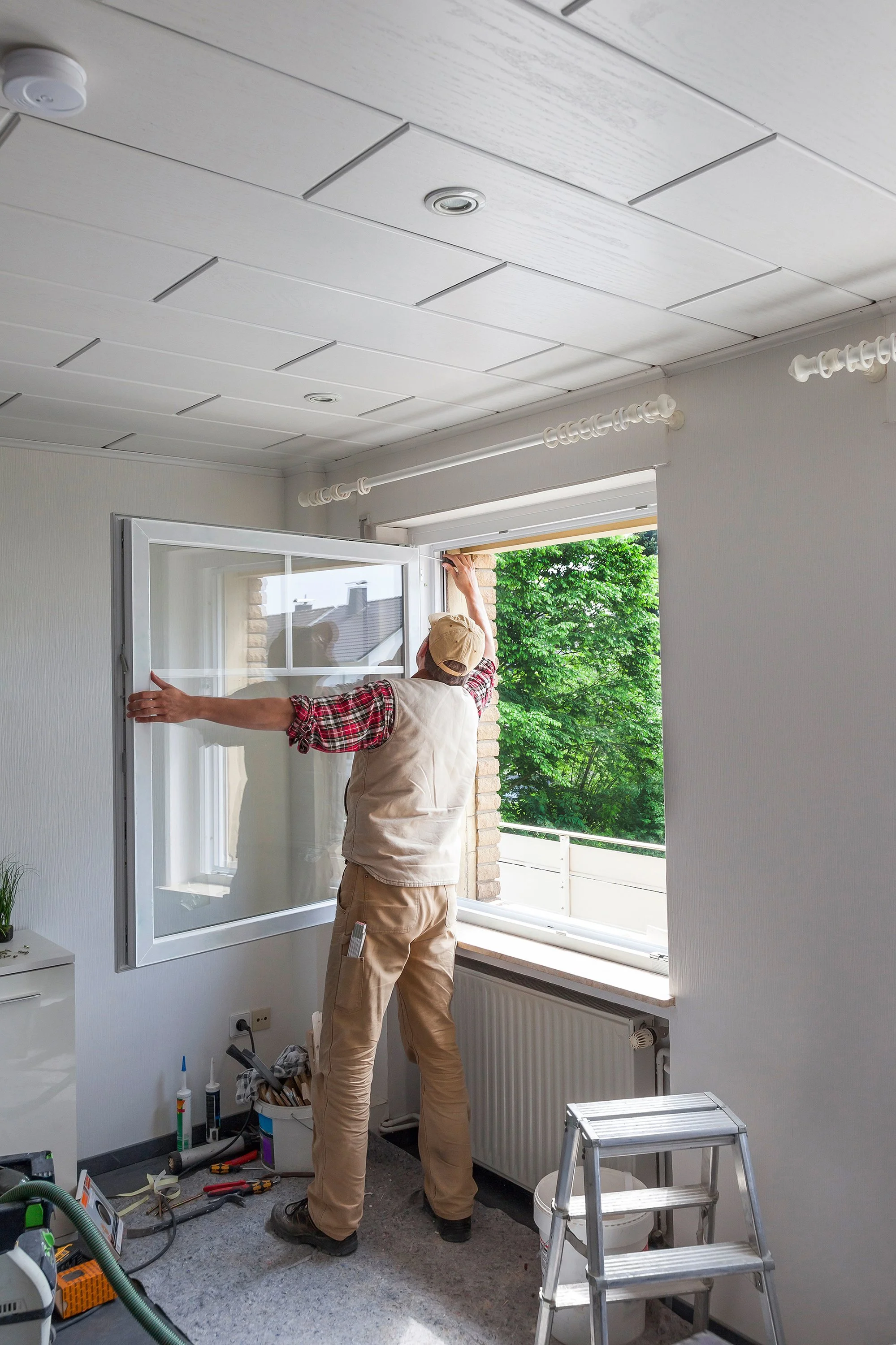 A man installing a window in a room with unfinished walls and building tools scattered on the floor.