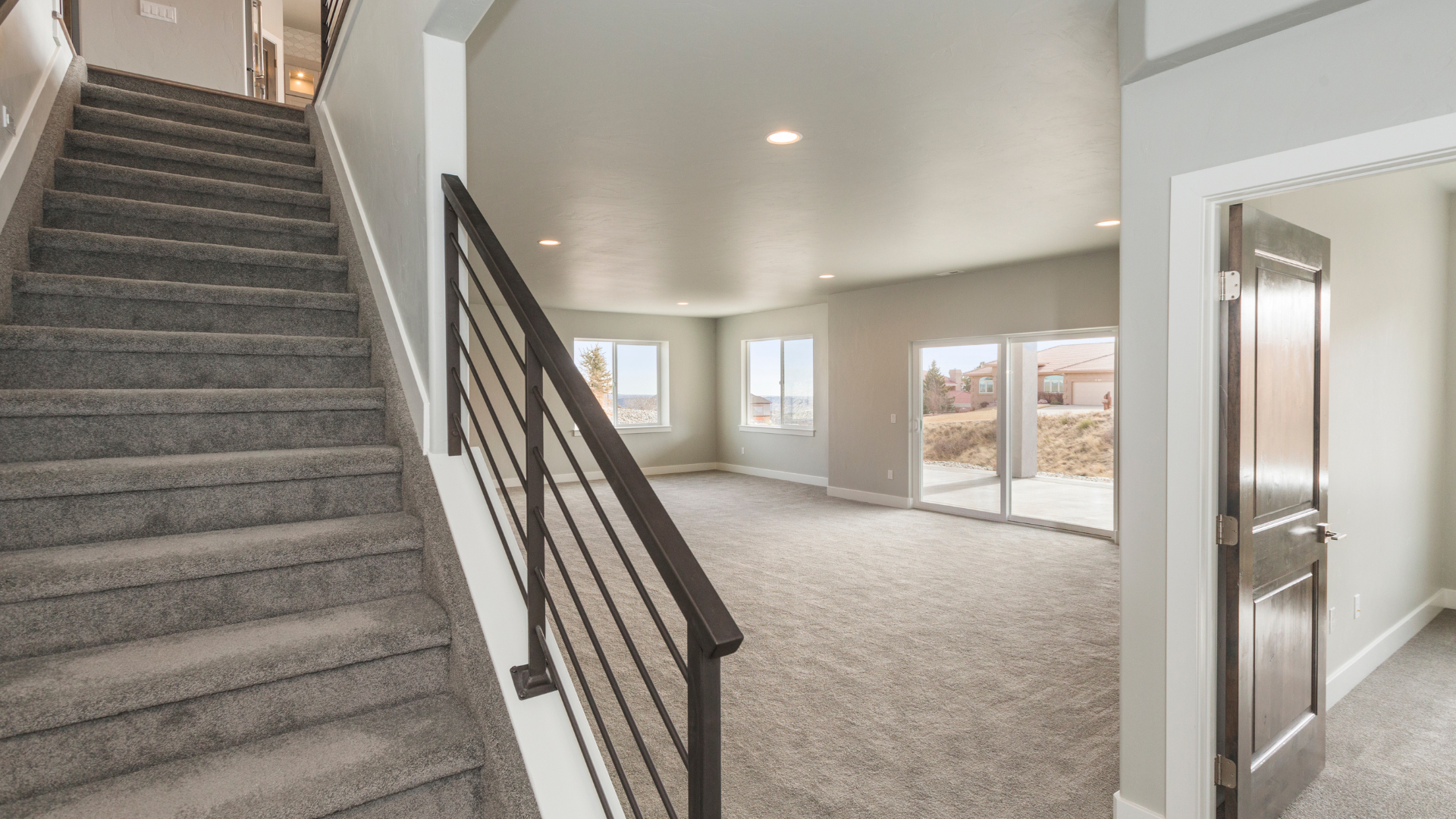 Modern house interior with carpeted stairs, large windows, glass sliding door, and neutral-colored walls, illuminated by recessed ceiling lights.