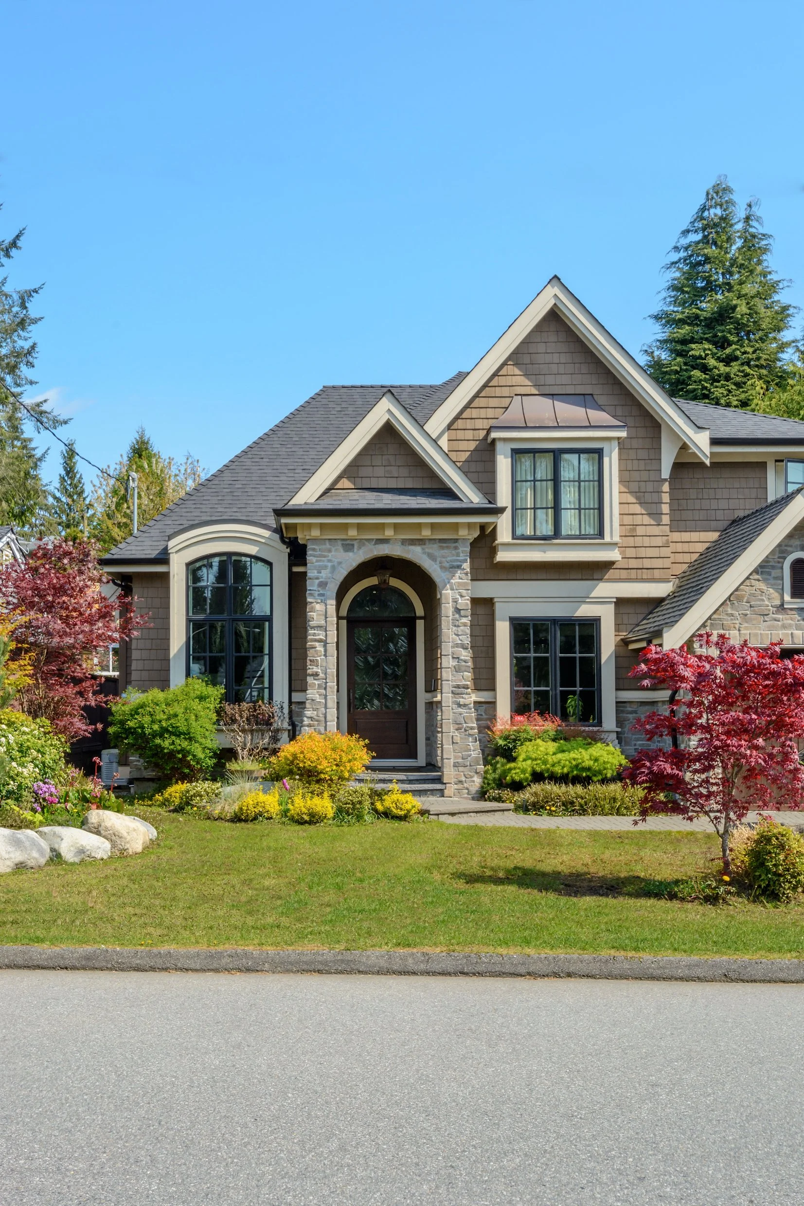 A two-story house with beige siding, stone accents, and dark gray roof, surrounded by a well-maintained garden with colorful shrubs and trees, and a gray street in the foreground.