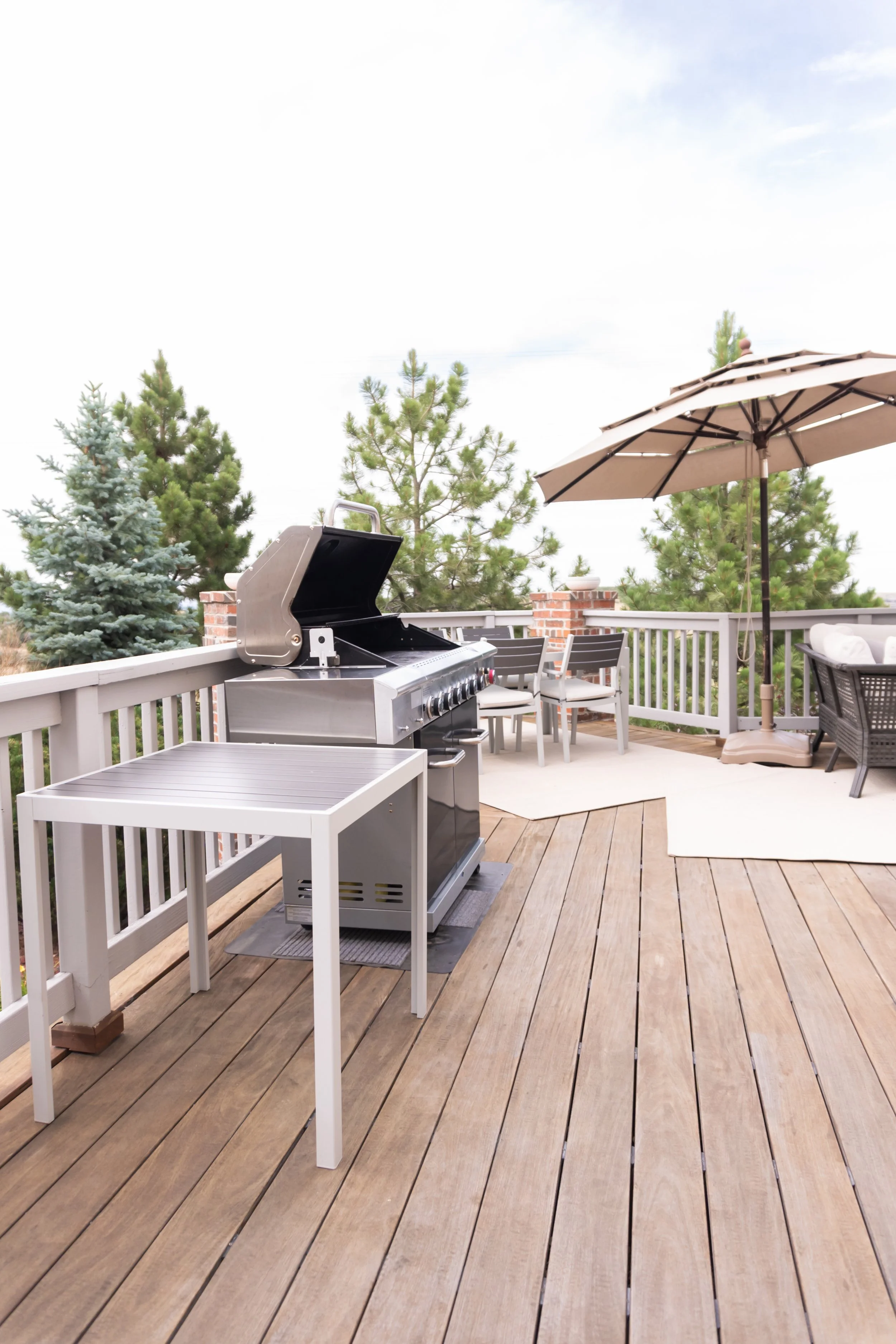 A spacious outdoor balcony with wooden flooring, a stainless steel grill, a small white table, an outdoor dining set with white chairs, a large beige patio umbrella, surrounded by pine trees, and a cloudy sky overhead.