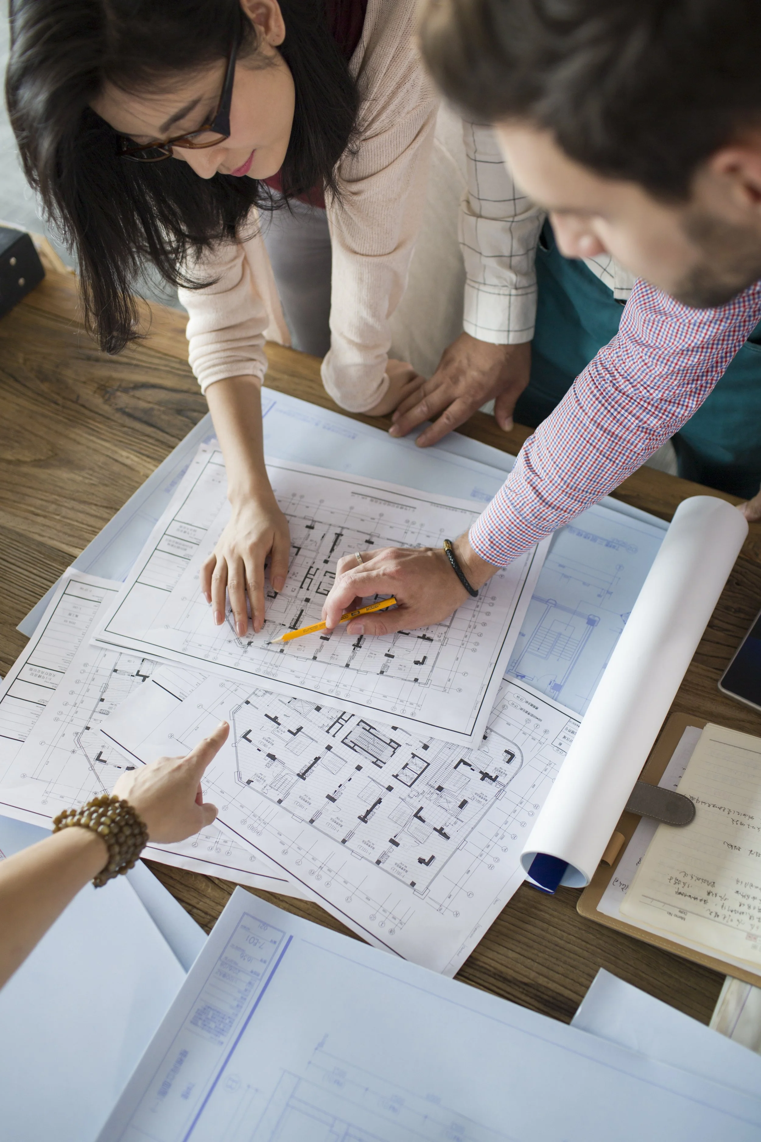 Two architects or engineers are reviewing detailed building floor plans spread out on a wooden table, pointing at specific sections.