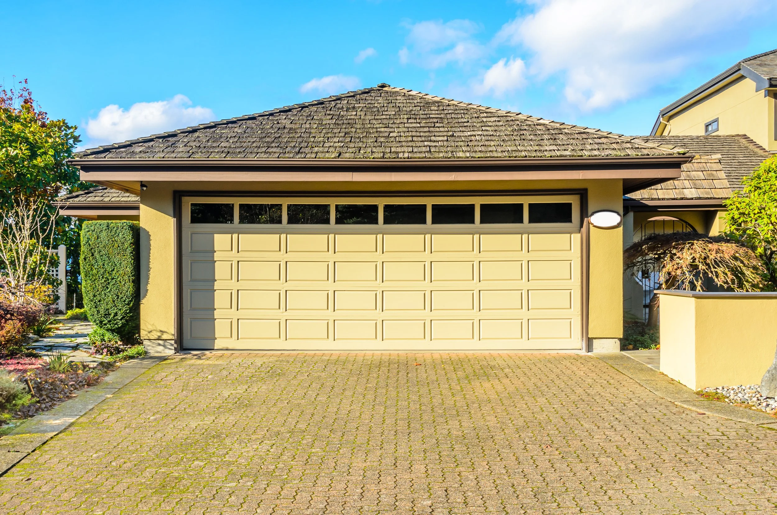 Front view of a house with a yellow garage door, tiled roof, and surrounding greenery under a blue sky with white clouds.
