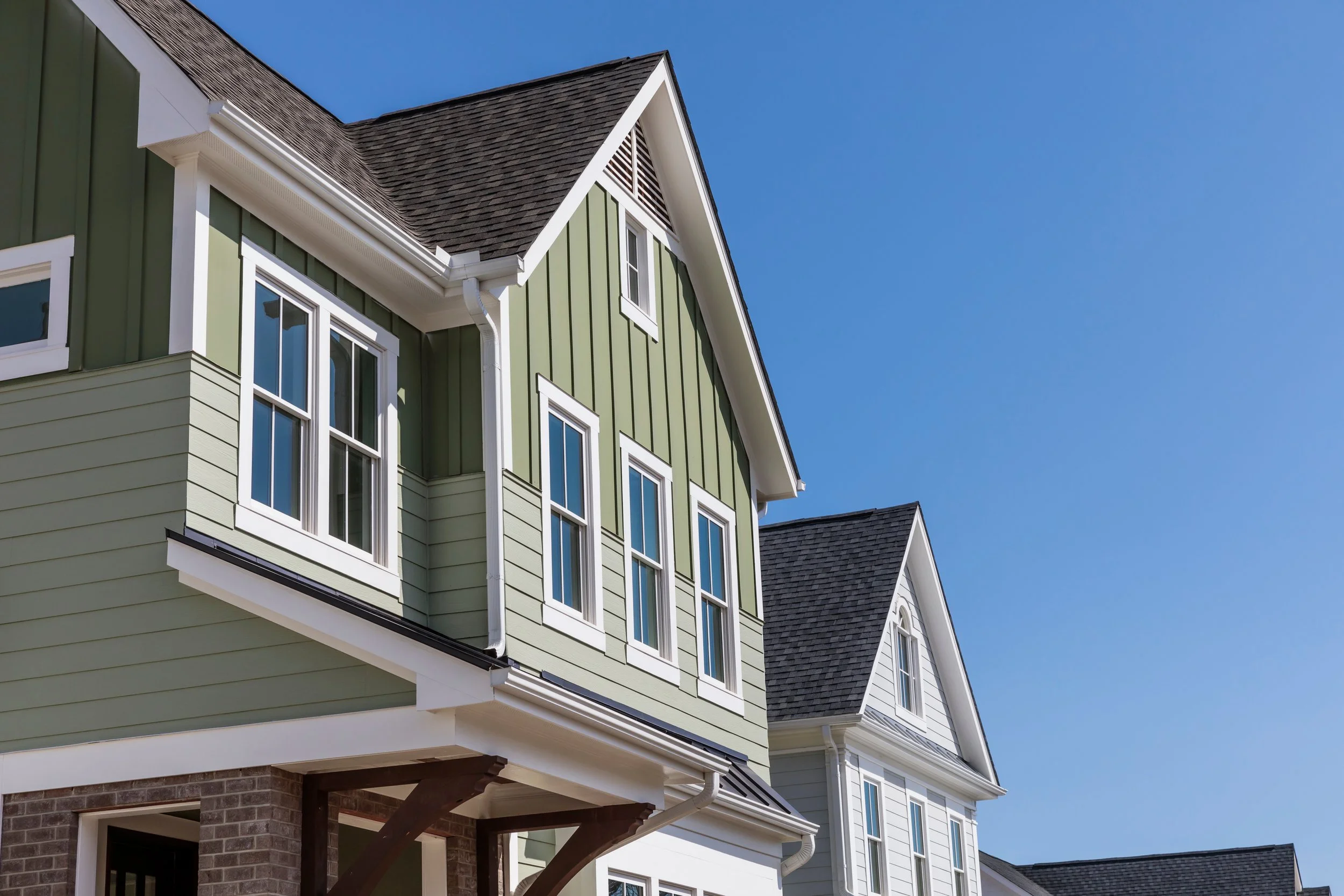 Modern multi-story houses with green and white siding, large windows, and dark gray shingle roofs under a clear blue sky.
