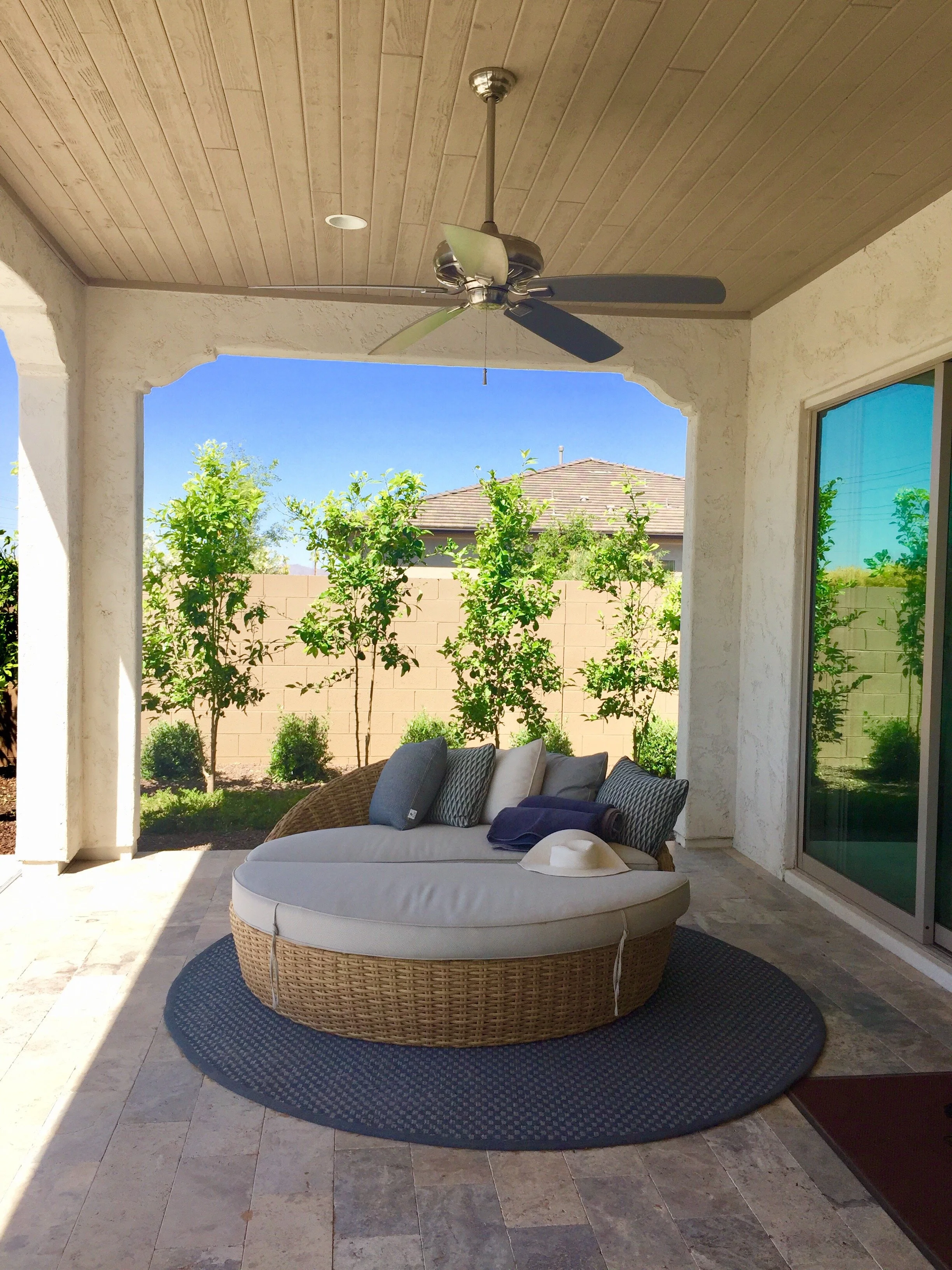 A covered patio area with a round cushioned wicker outdoor sofa set on a black circular rug. There is a white hat and a rolled-up towel on the sofa, with a background of green trees and a brick wall, under a ceiling fan with five blades.