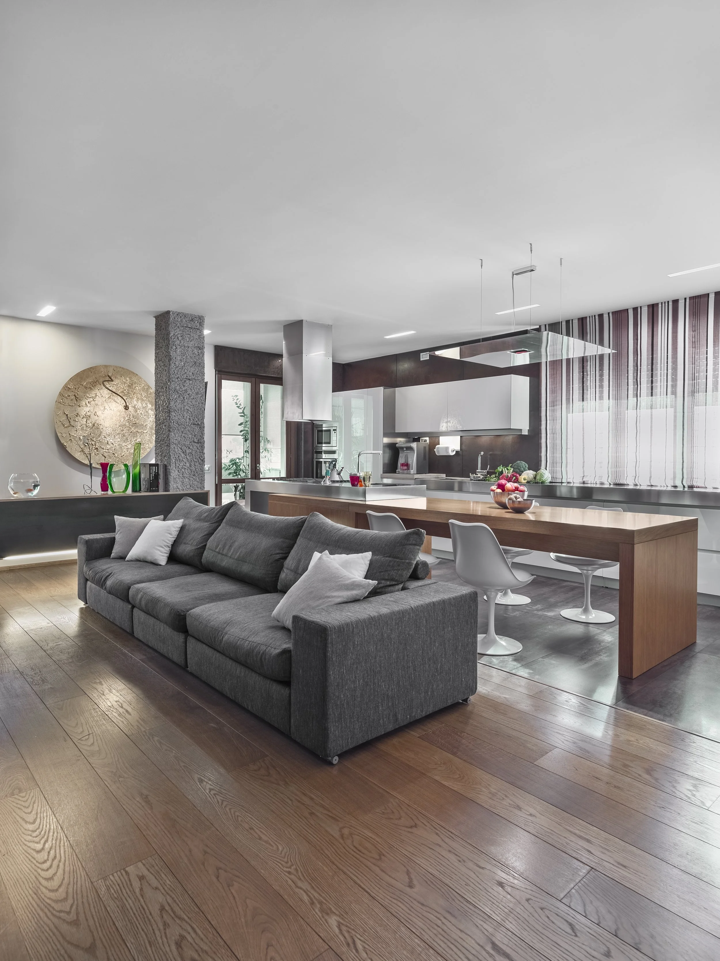 Open-concept living room and kitchen with hardwood floors, gray sofa with pillows, modern dining table with white chairs, kitchen with white cabinets, stainless steel appliances, and a large clock on the wall.