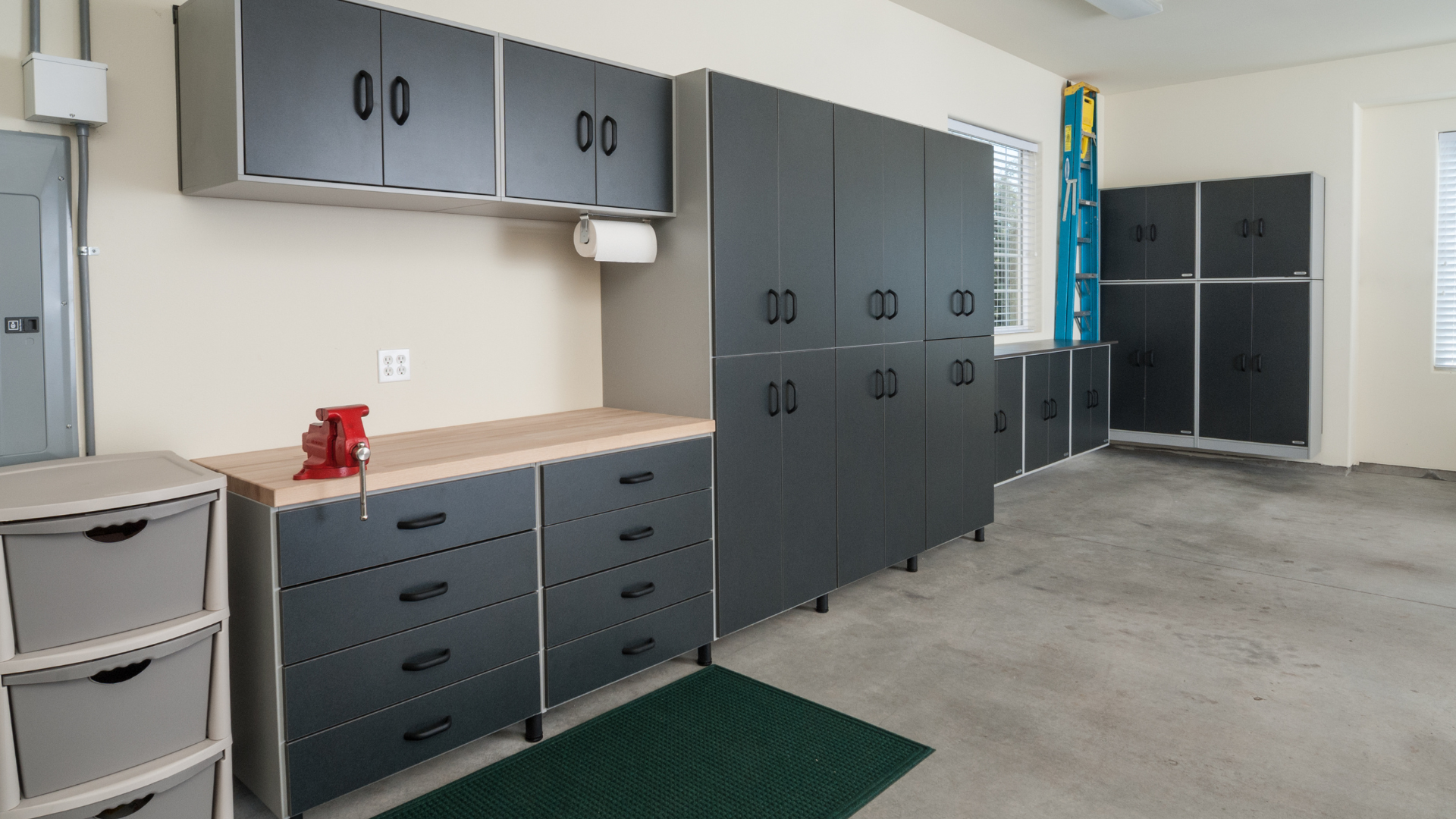 Clean garage with gray cabinets, a workbench with a red clamp, plastic storage drawers, a ladder, and windows with blinds.