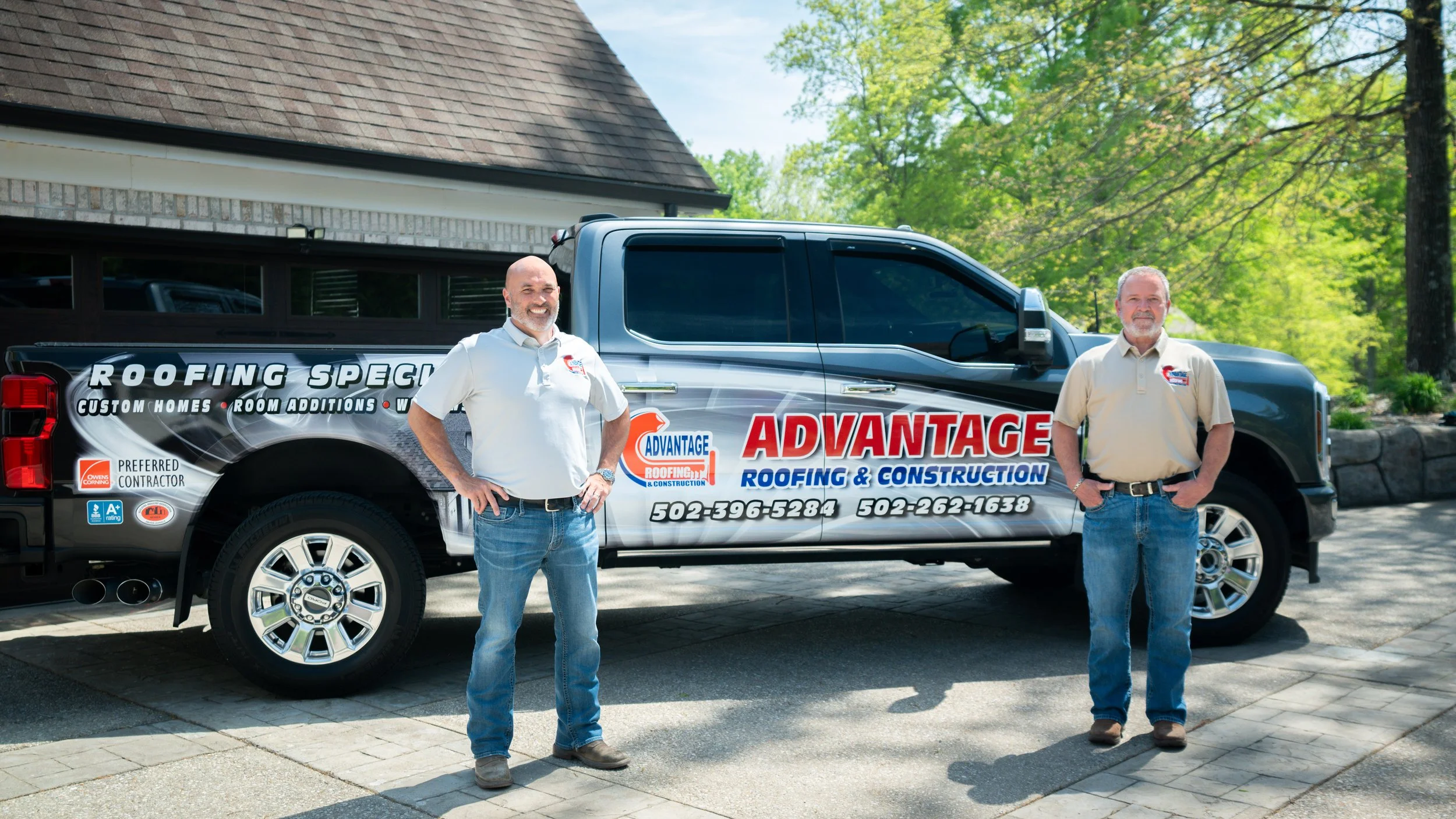 Two men standing in front of a truck with company branding for roofing and construction, outdoors with green trees and a house in the background.