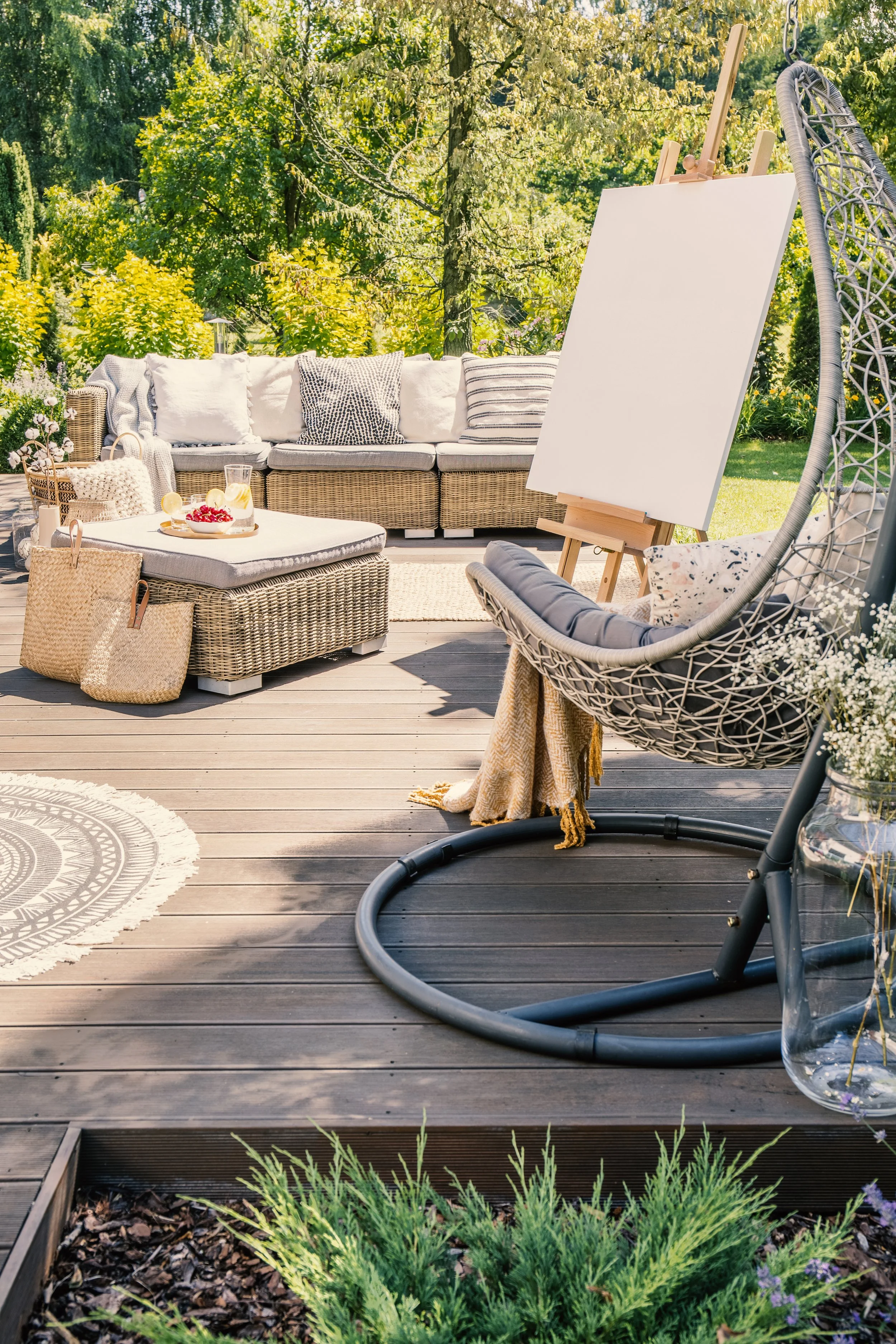 An outdoor patio scene with a wicker sofa with white cushions and decorative pillows, a matching ottoman with a tray of refreshments and cherries, a wicker basket, a standing easel with a blank canvas, a hanging egg chair with cushions, and a jar with flowers on a wooden deck surrounded by greenery and plants in a garden.