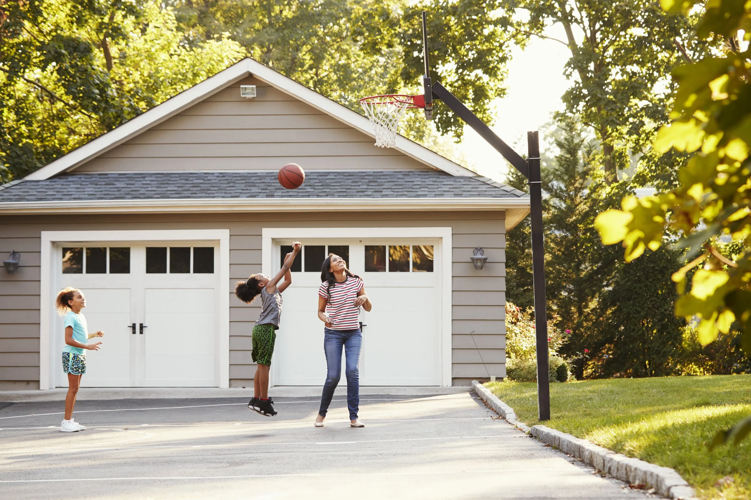A woman and children playing basketball outside a house, with a basketball hoop mounted on the garage, during sunset.