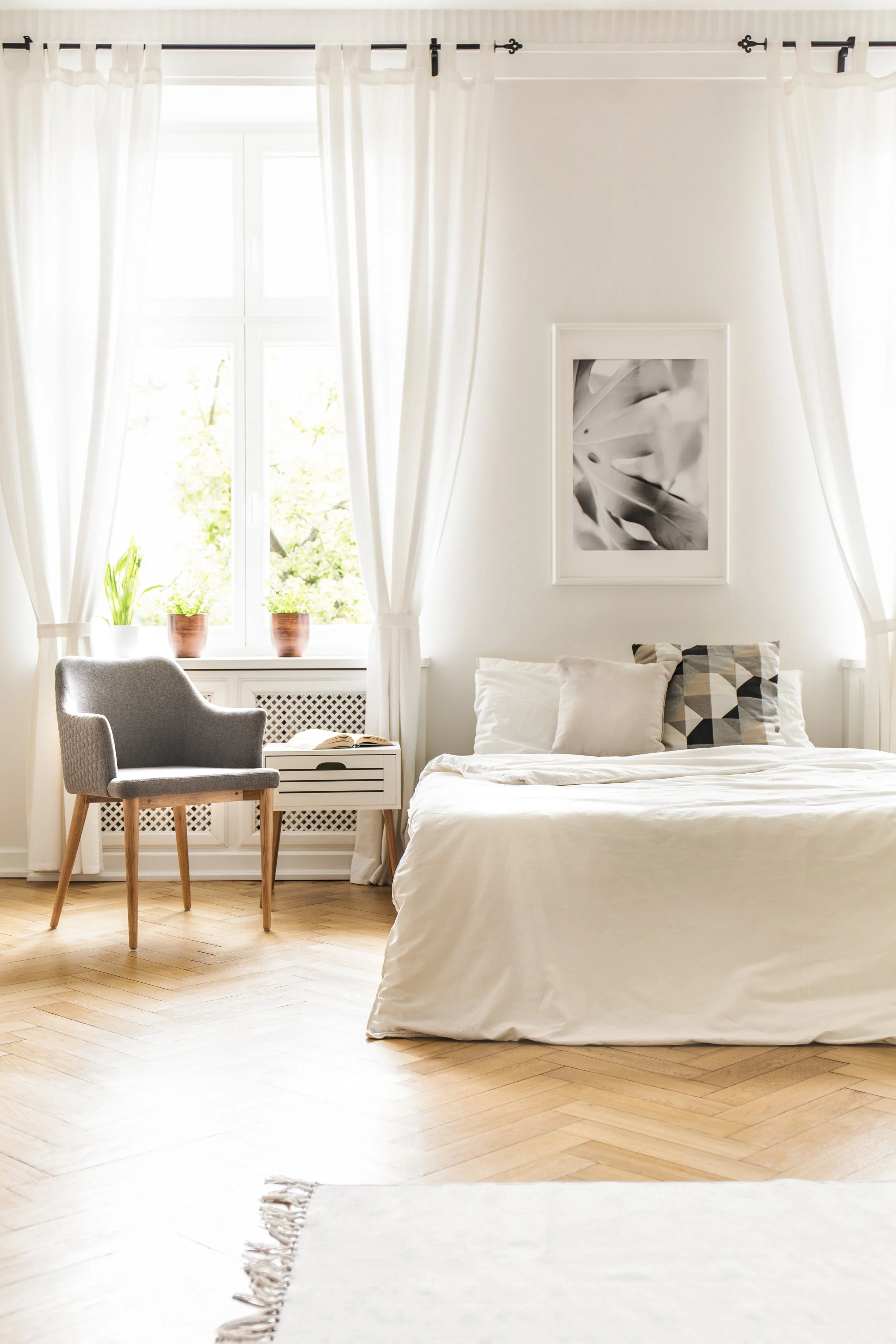 Bright bedroom with large window, white curtains, green plants on windowsill, gray armchair facing window, white nightstand, bed with white bedspread, decorative pillows, framed artwork on wall.