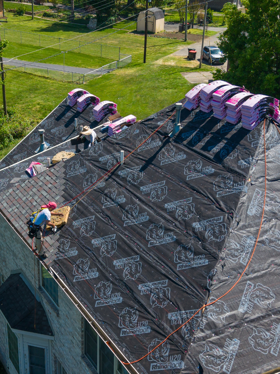 Roofers installing black roofing underlayment on a house roof with multiple stacks of shingles, overlooking a backyard with a tennis court, a shed, and a parked car.