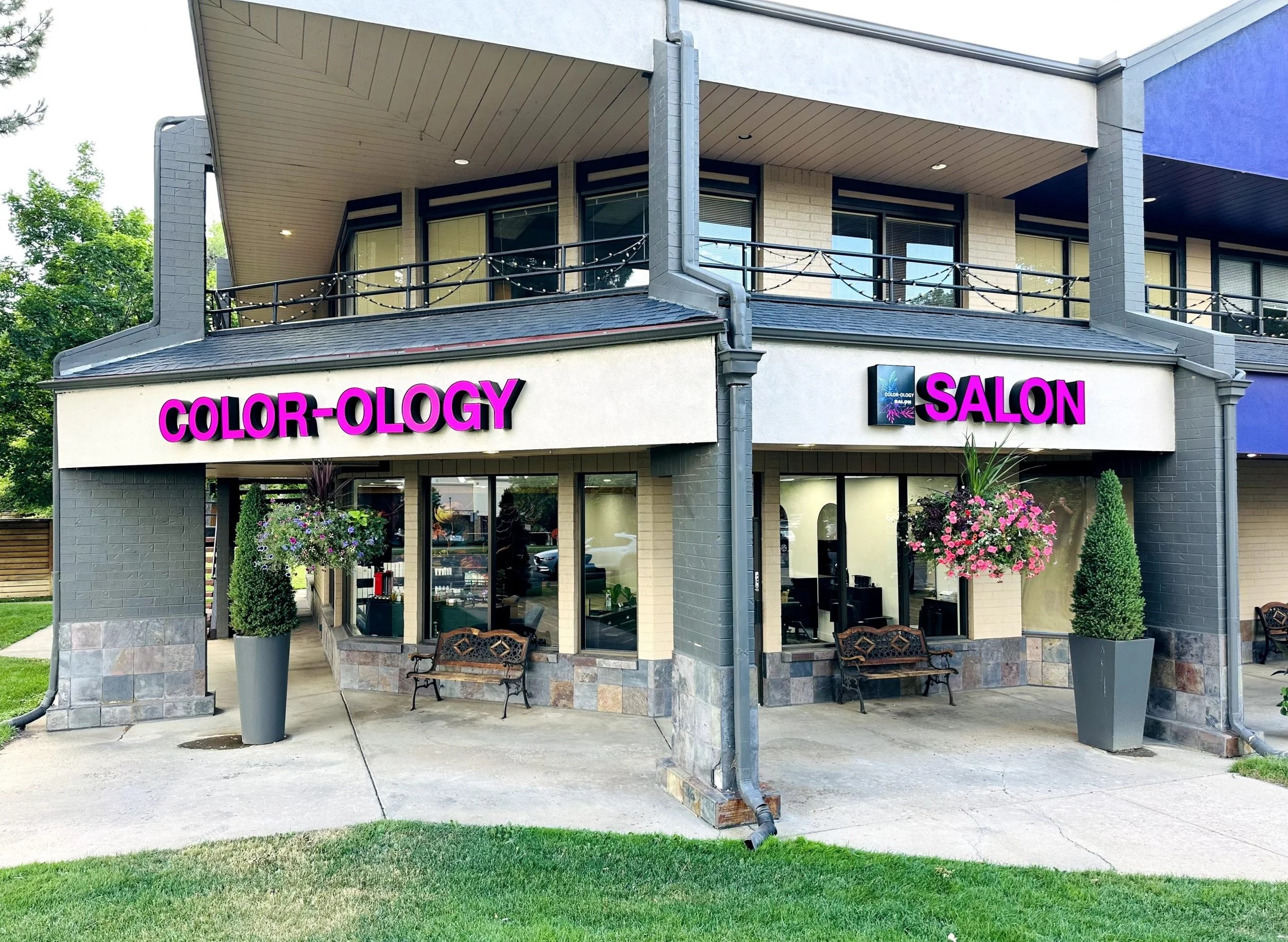 Exterior of a salon with a sign that reads 'Color-ology' in purple letters and 'Salon'. The salon has large windows, potted plants, hanging flower baskets, and benches outside.