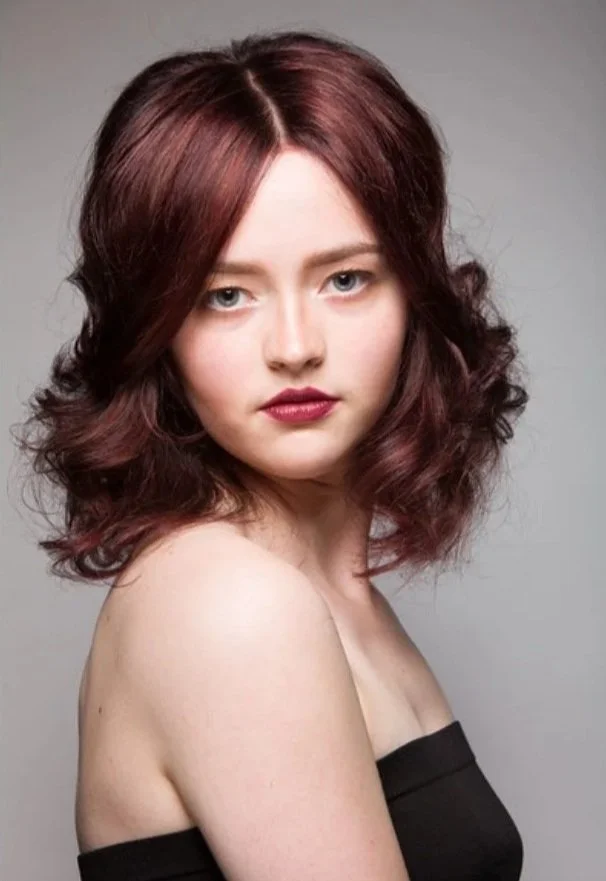 A young woman with wavy, shoulder-length reddish-brown hair looking at the camera with a neutral expression, wearing a black off-the-shoulder top against a plain gray background.