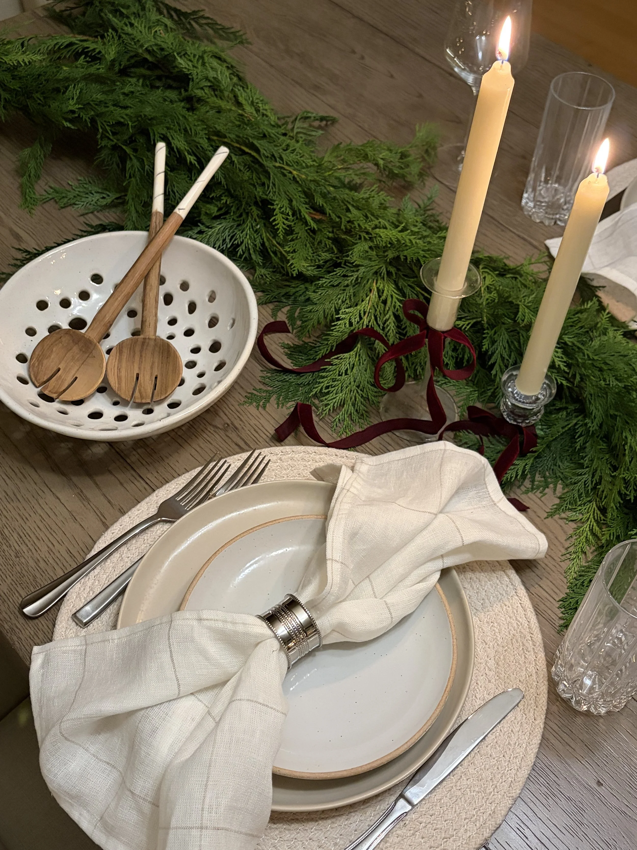 A holiday table setting with cream-colored plates, a beige napkin tied with a silver napkin ring, silverware, lit candles, and greenery with candles in glass holders for decoration.