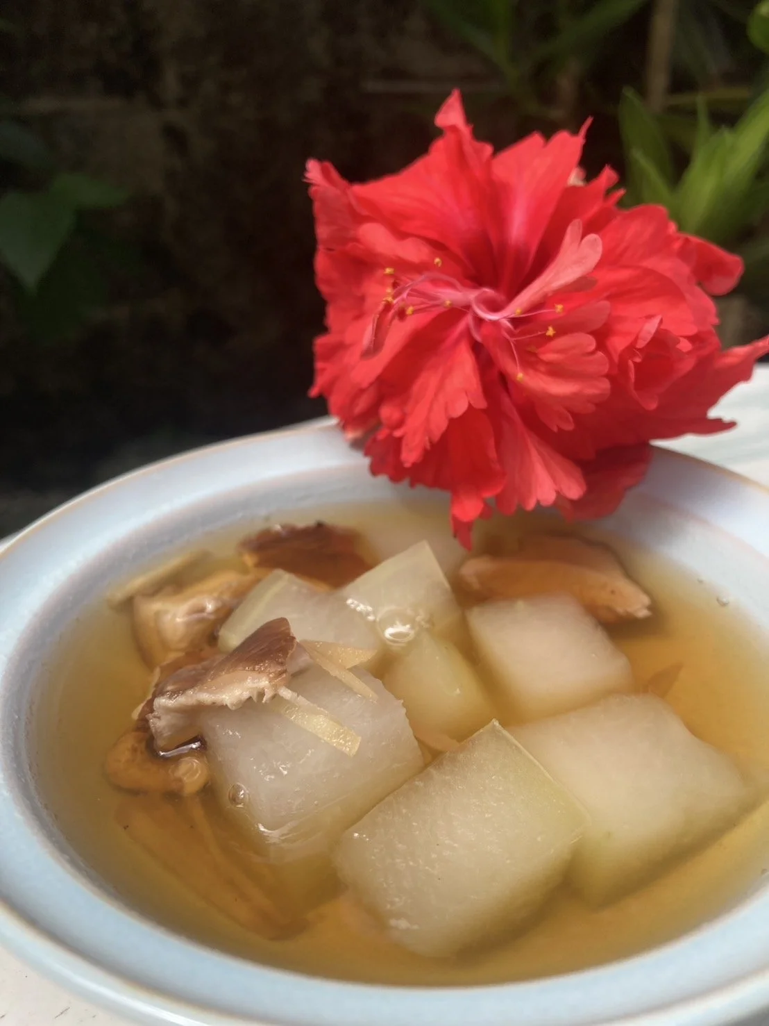 A bowl of clear broth with chunks of white radish and mushrooms, garnished with a red hibiscus flower.