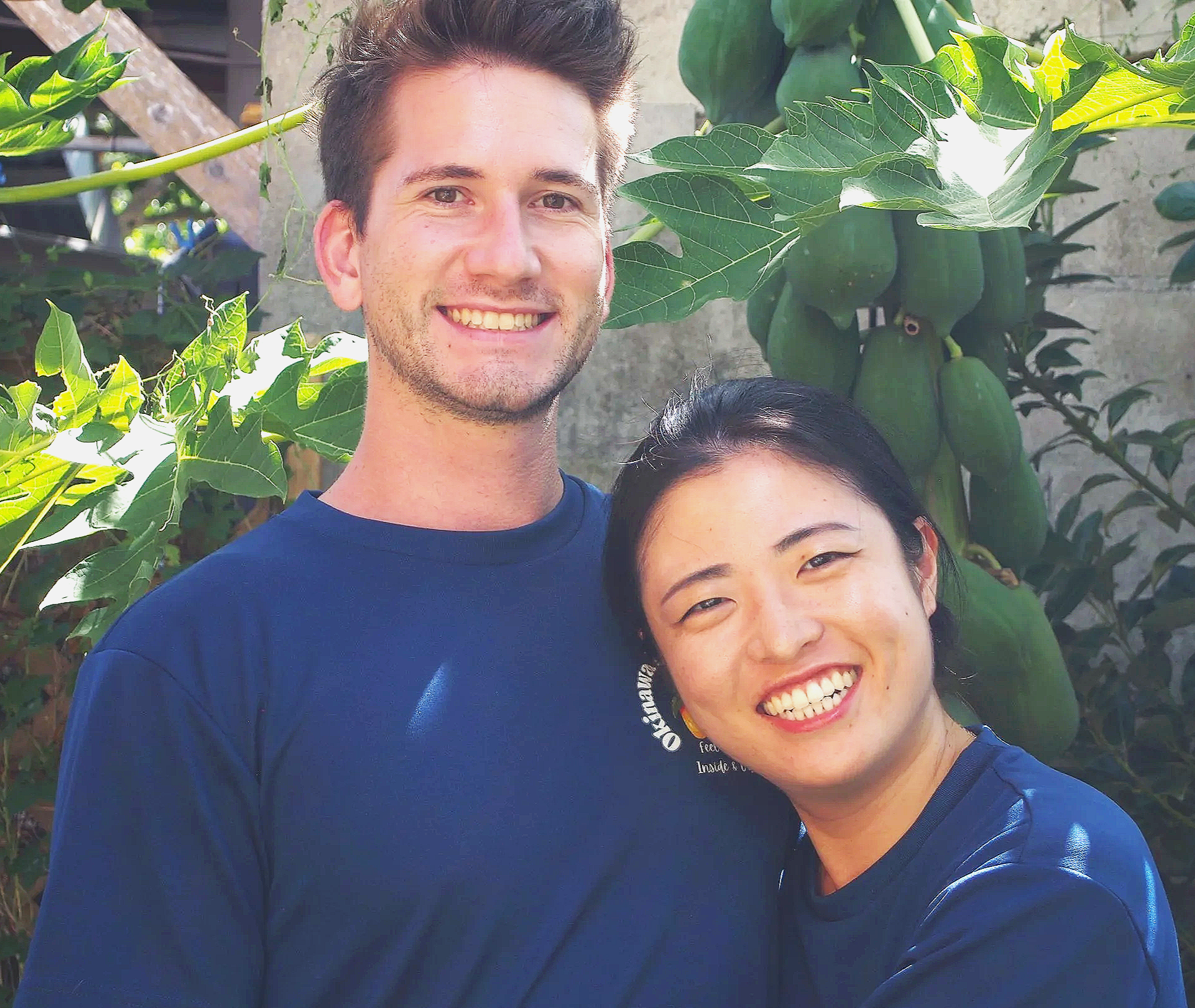 Two smiling individuals, a man and a woman, standing outdoors in front of a leafy plant with green fruits. They are both wearing dark blue shirts and appear happy.