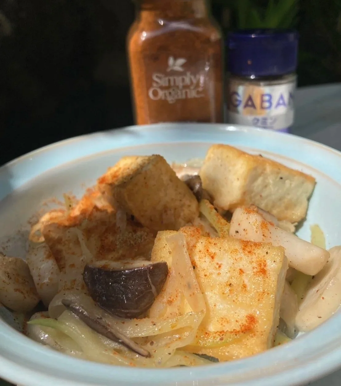 Close-up of a bowl containing fried tofu, onions, and black mushrooms, seasoned with paprika. In the background, there are bottles of organic syrup and ginseng extract.
