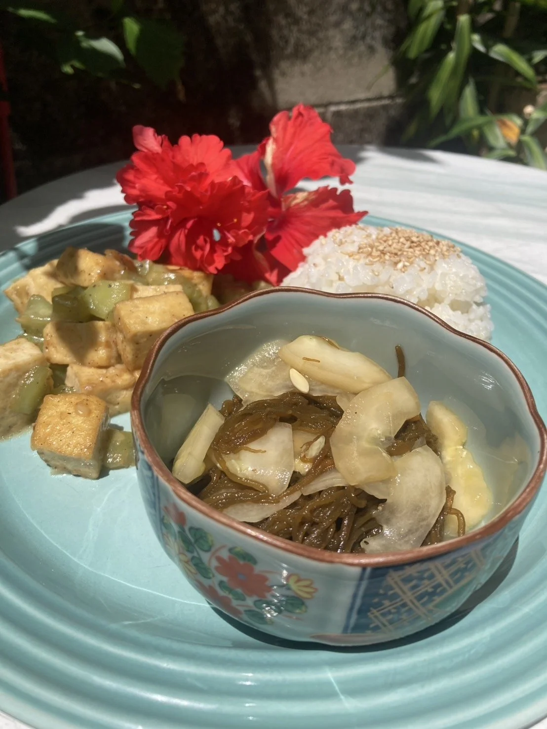 A plate of Asian-inspired food featuring rice topped with sesame seeds, a vegetable stir-fry with tofu, seaweed salad with onions, and a red flower for decoration outdoors.