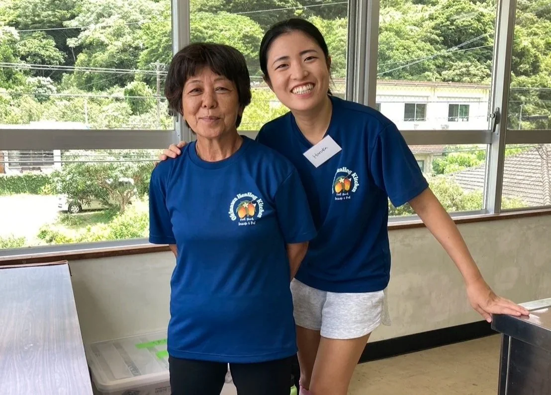 Two women smiling, wearing matching blue shirts with a logo, inside a room with large windows showing trees and houses outside.