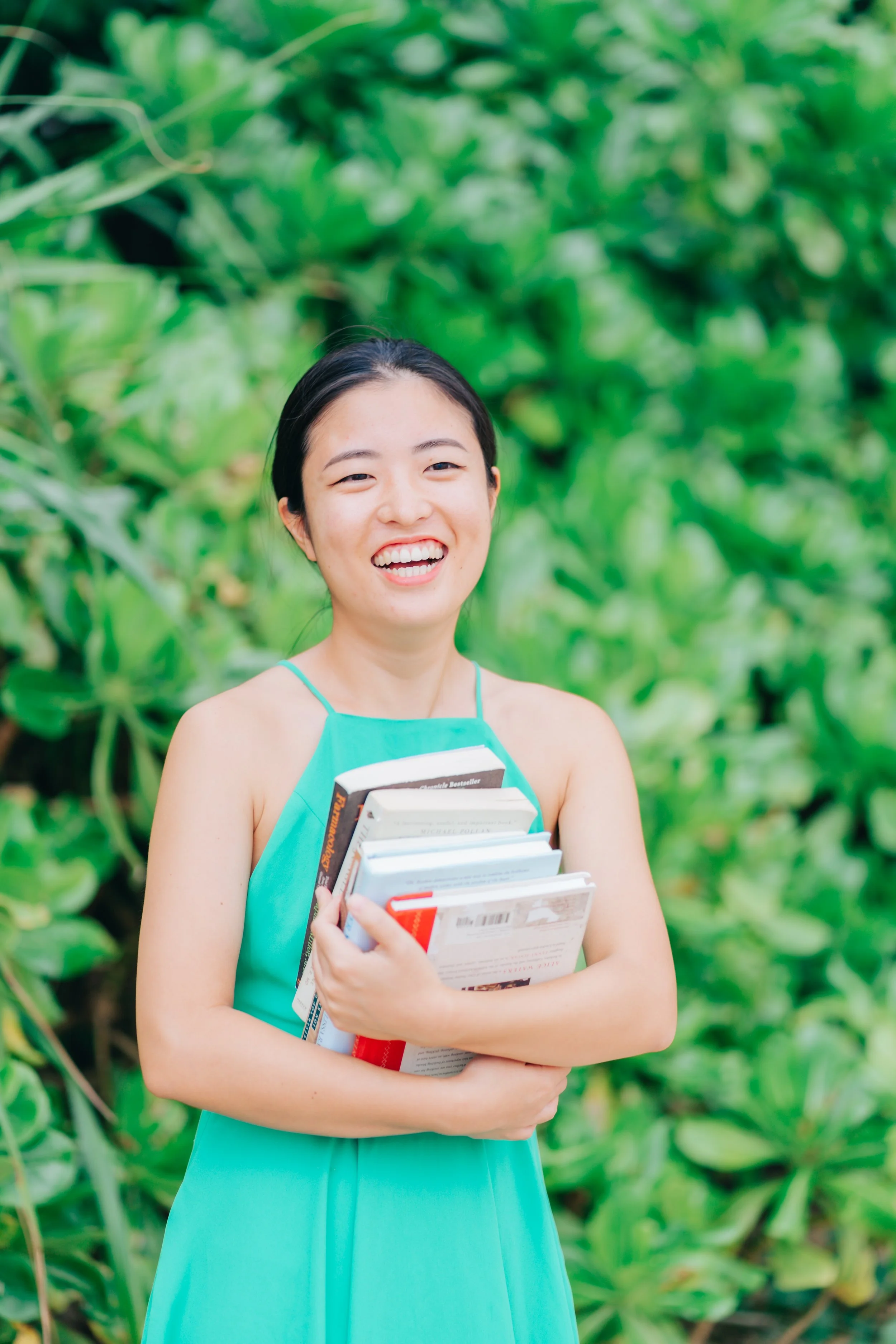 A young woman with black hair tied back, smiling and holding several books, standing outdoors with lush green foliage in the background. She is wearing a sleeveless turquoise dress and appears happy and relaxed.