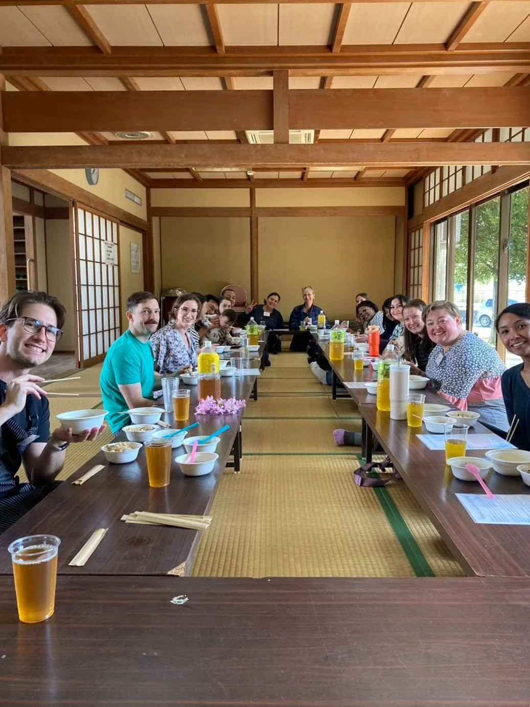 Group of people seated around traditional Japanese low tables with tatami mats, enjoying a meal in a Japanese-style room with sliding shoji doors, windows, and wooden beams.