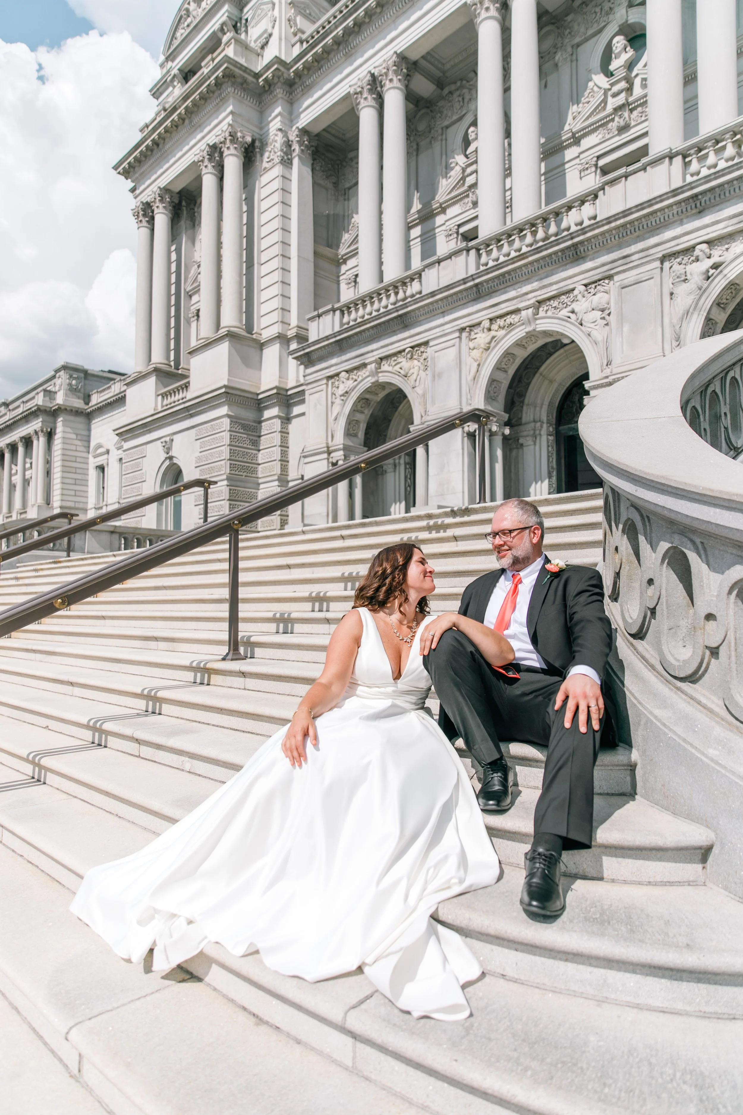 A bride and groom sitting on the steps of a grand, historic building with classical architecture, including columns and ornate details, on a cloudy day.