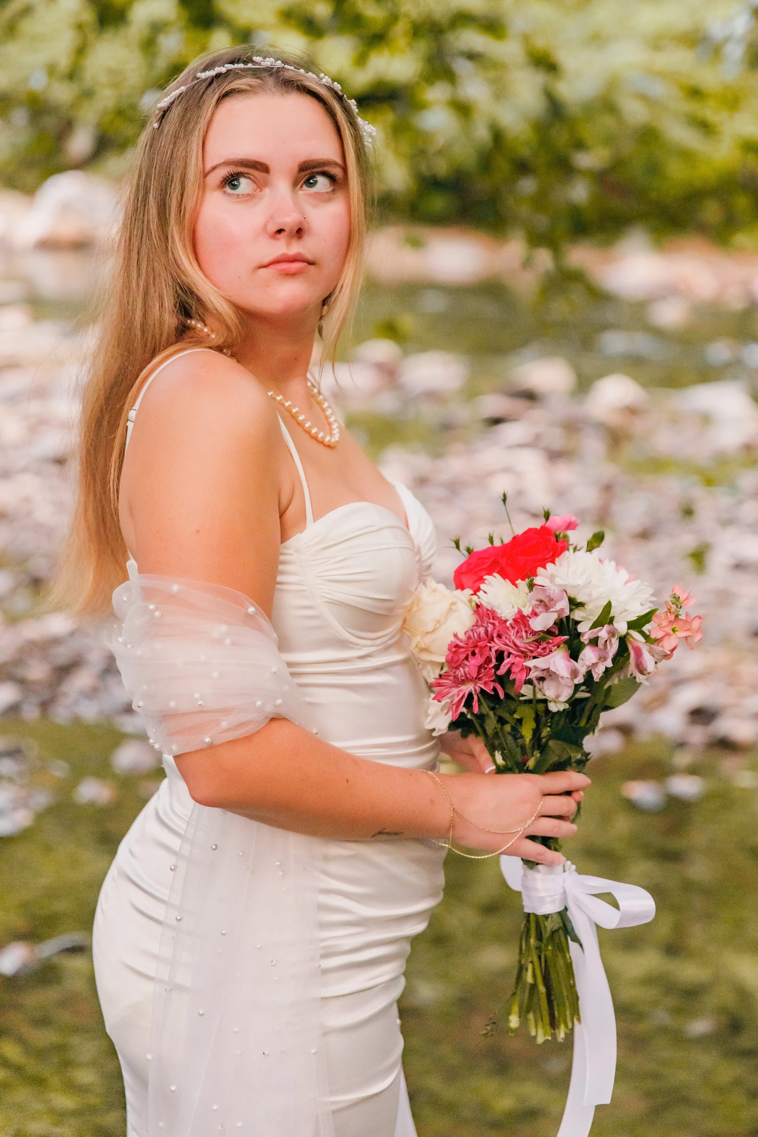A woman in a white wedding dress holding a bouquet of pink, red, and white flowers, standing outdoors near a body of water with greenery in the background.