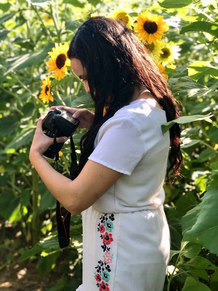 A woman with long dark hair wearing a white dress with colorful embroidery on the side, holding a camera and looking down at it in a sunflower field.
