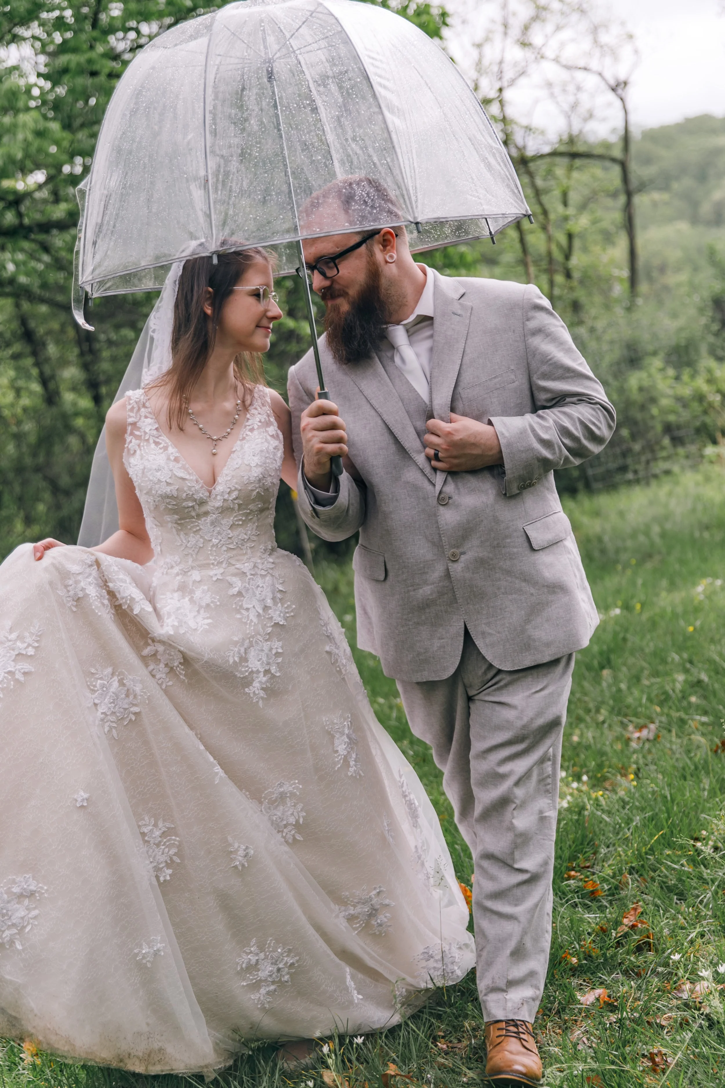 A couple walking through a field with an umbrella on their elopement day smiling at each other