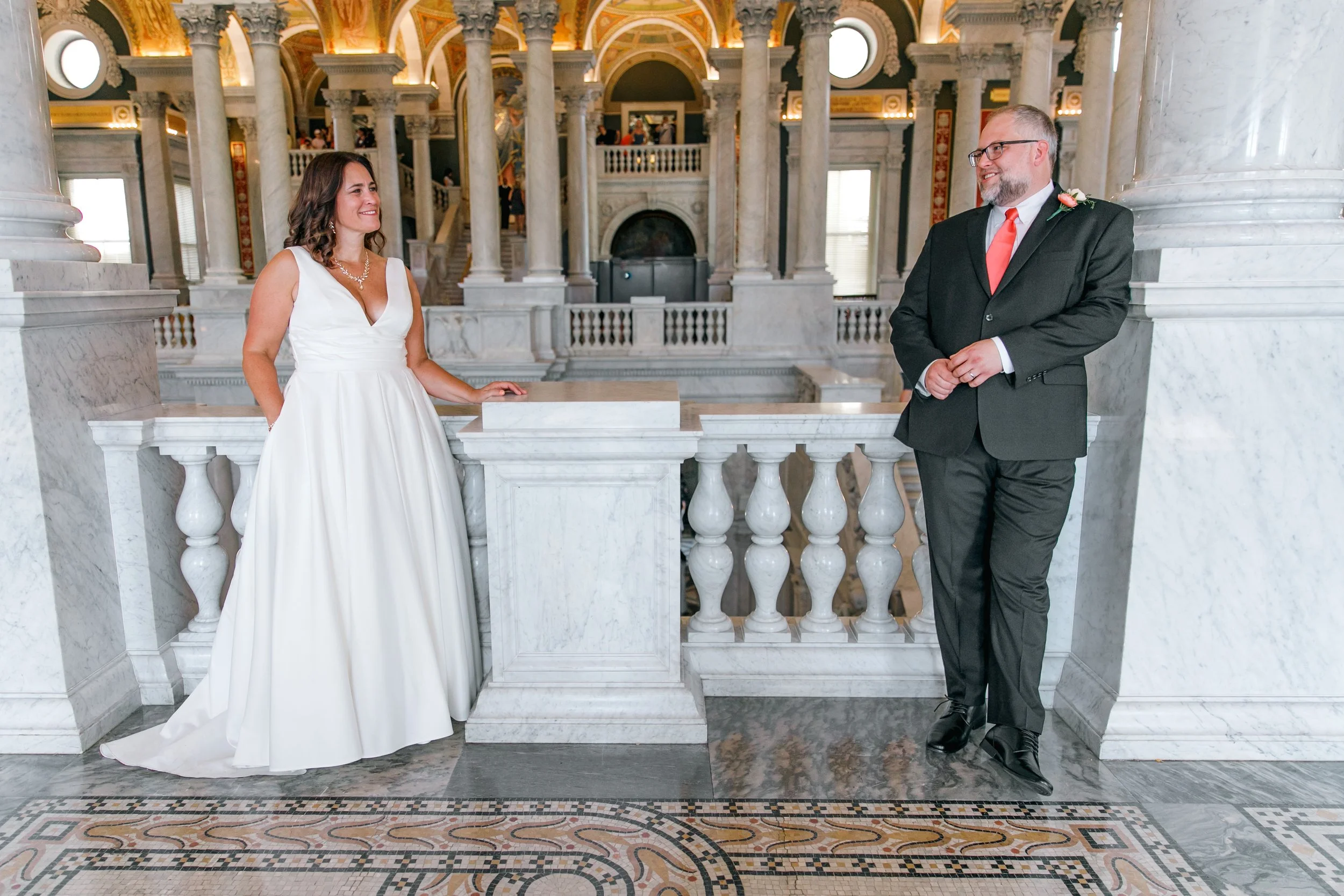 A bride and groom stand facing each other inside a grand marble hall. The bride wears a white gown and the groom is dressed in a dark suit with a red tie. They are smiling at each other near a marble railing with classic architectural columns and a d